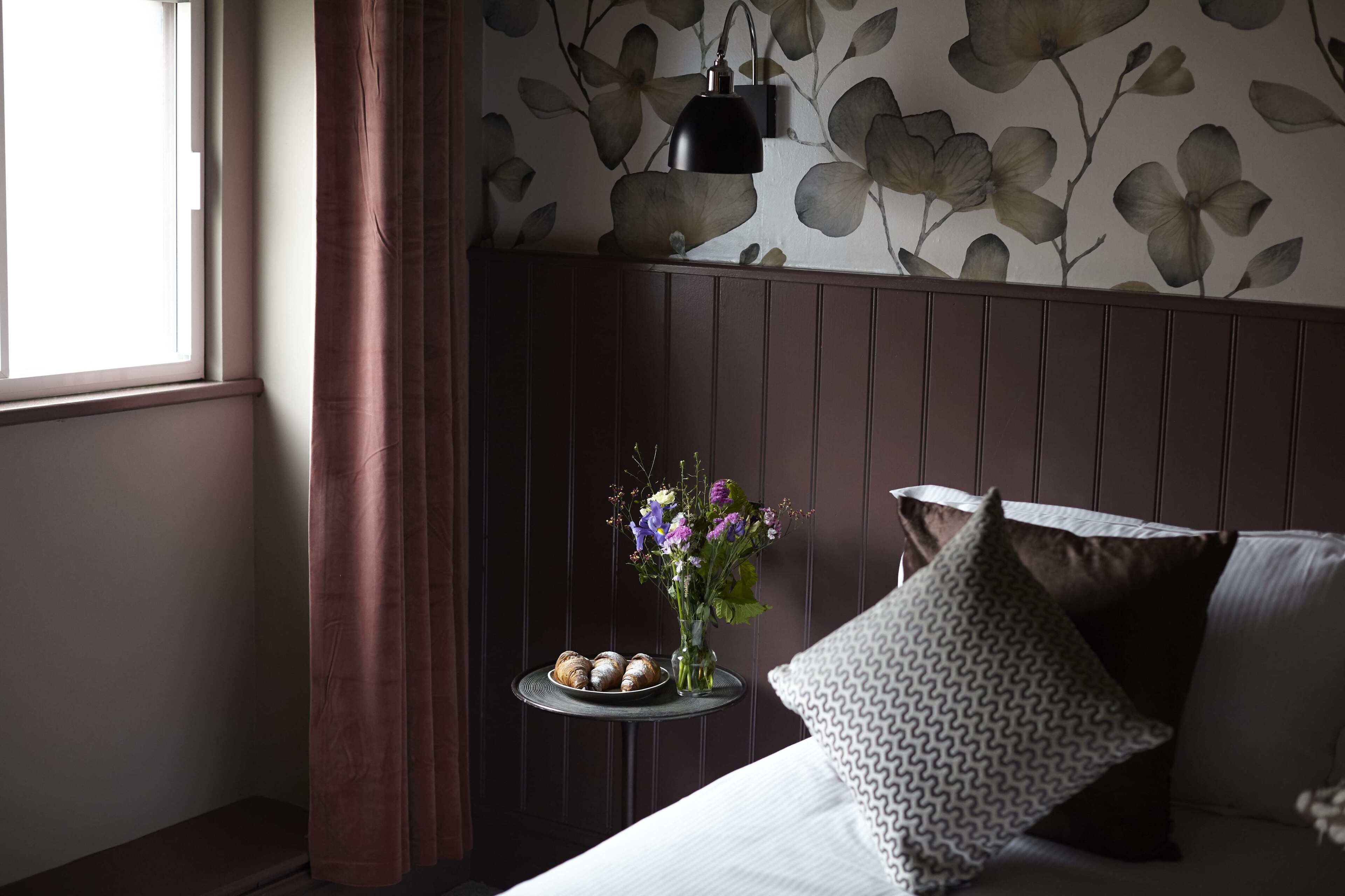 A bedroom with a bed, a bedside table with a vase of flowers and pastries, a wall with floral wallpaper, and a window with curtains.