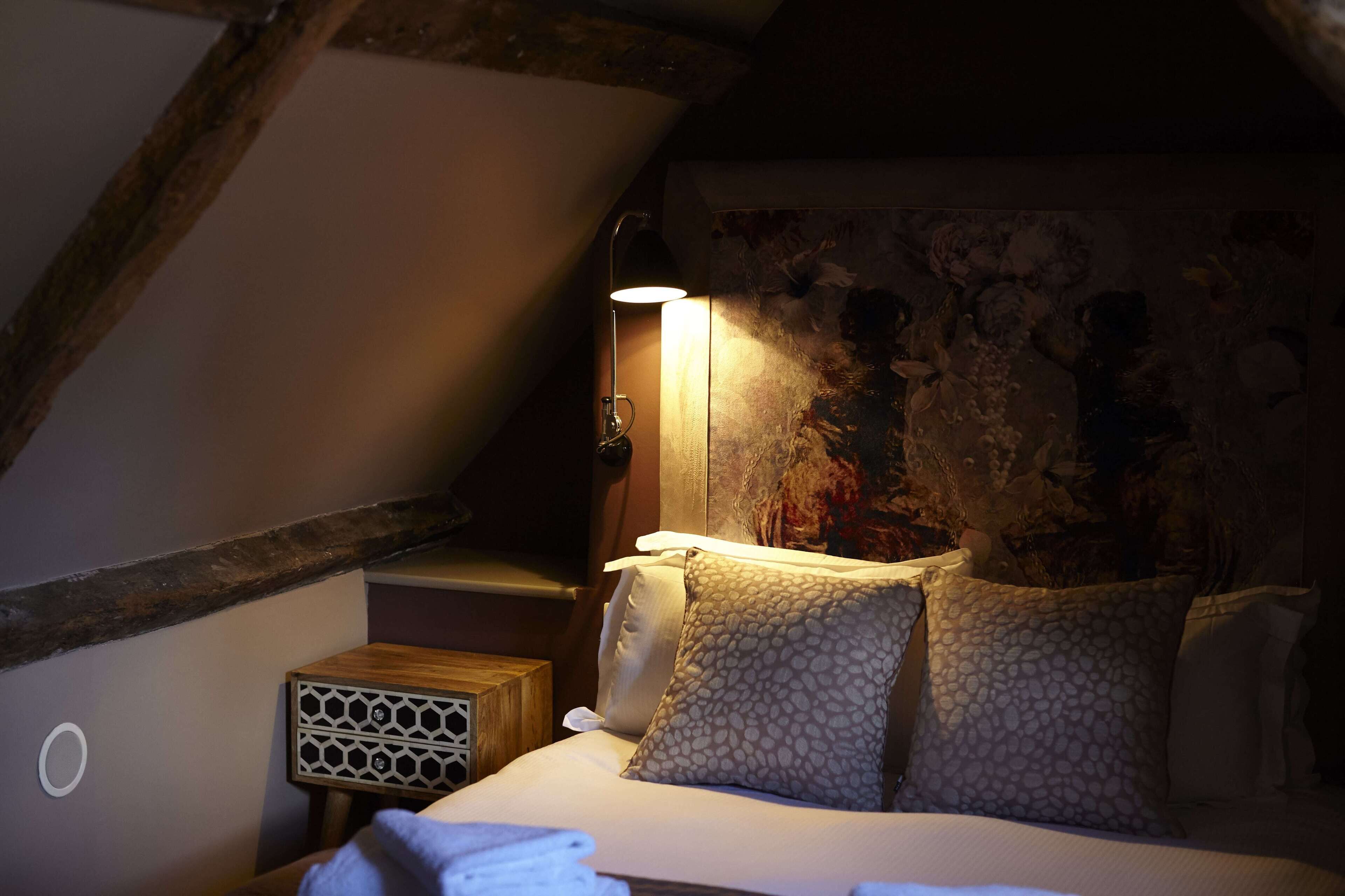 A cozy bedroom with a sloped ceiling, a bed with patterned pillows, a bedside lamp, and a small wooden nightstand.