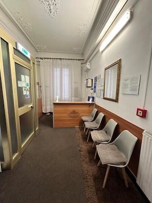 A reception area with a wooden desk, a waiting area with chairs, a door with a glass panel, and a wall with framed pictures and notices. - Hotel Barone (Genoa)