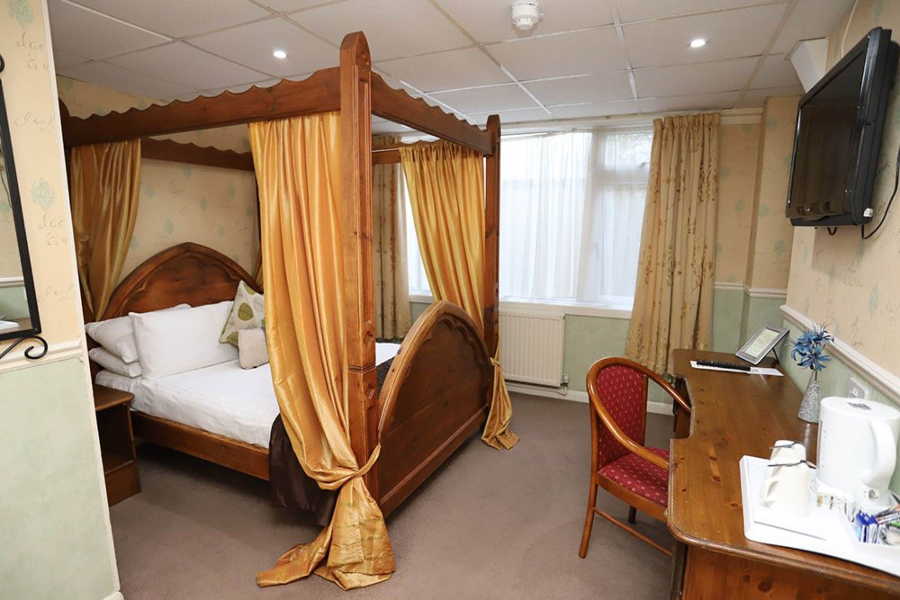 A four-poster bed with curtains, a wooden desk with a chair, a television, and a radiator.