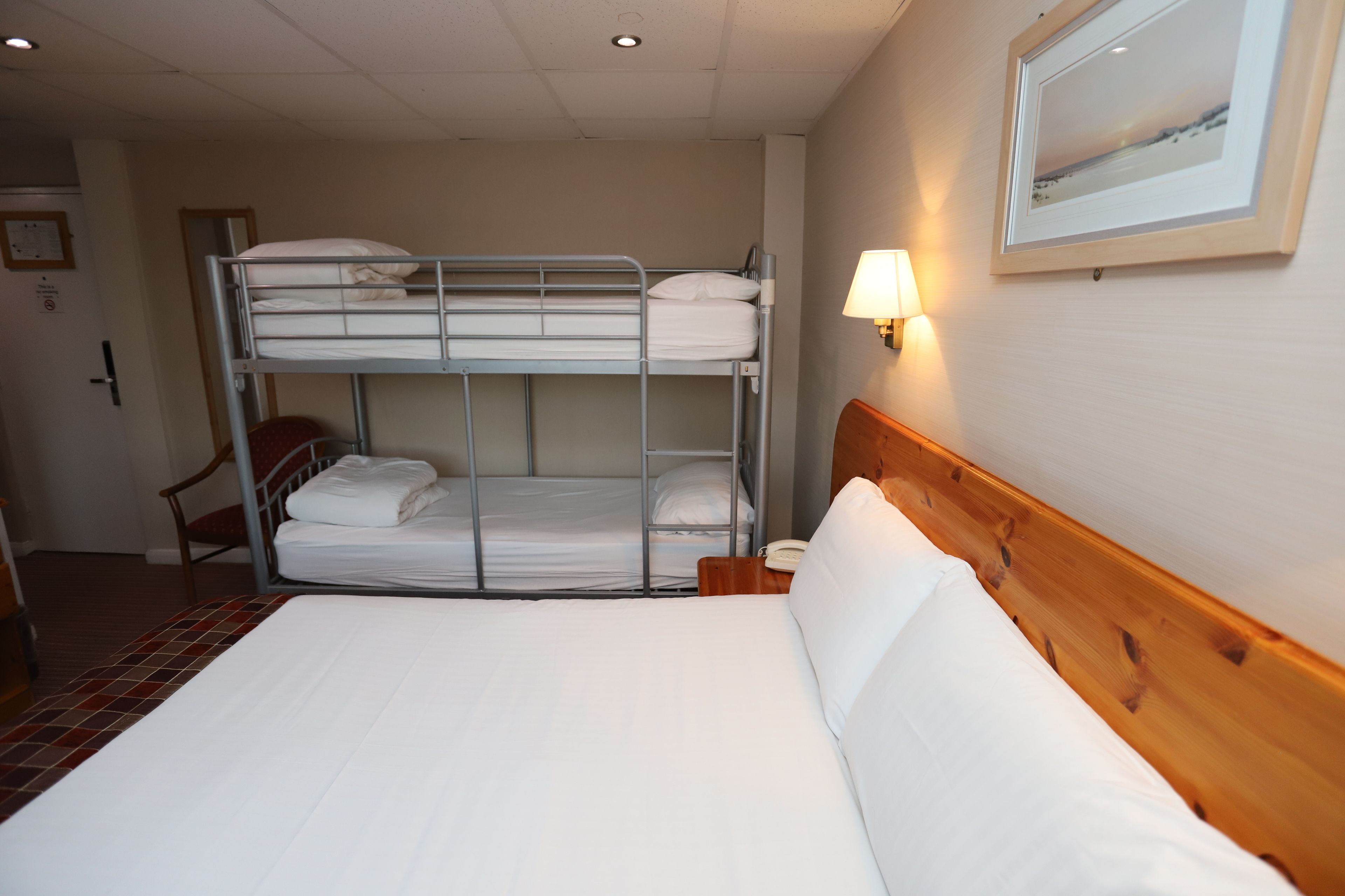 Habitación de hotel con literas, cama individual, cabecera de madera, lámpara de noche y un cuadro en la pared.