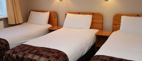 Une chambre d’hôtel avec trois lits simples, chacun doté d’une tête de lit en bois et de linge de lit blanc. Deux lampes murales assurent l’éclairage.