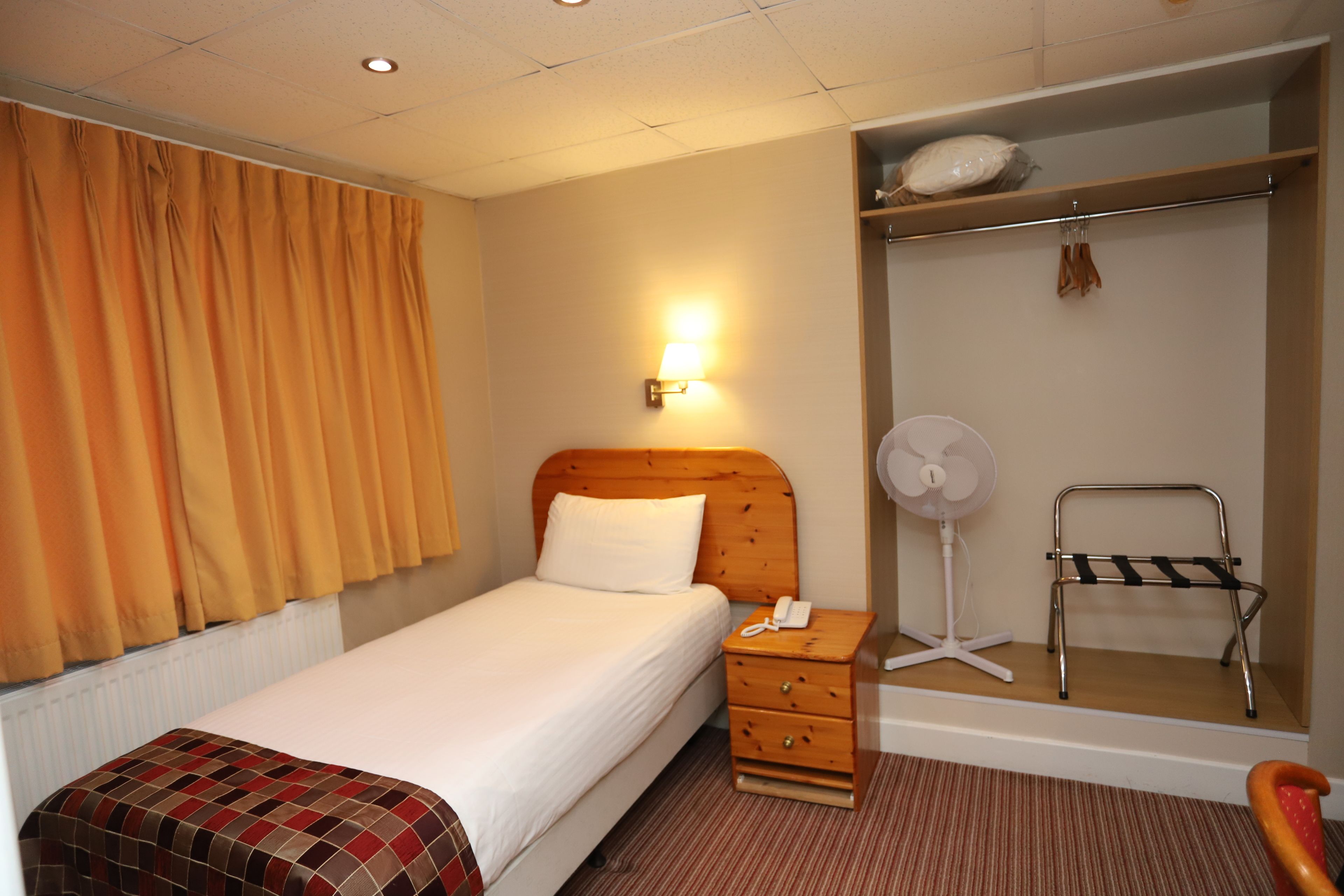 A hotel room with a bed, a bedside table with a lamp, a fan, a towel rack, and a wardrobe.