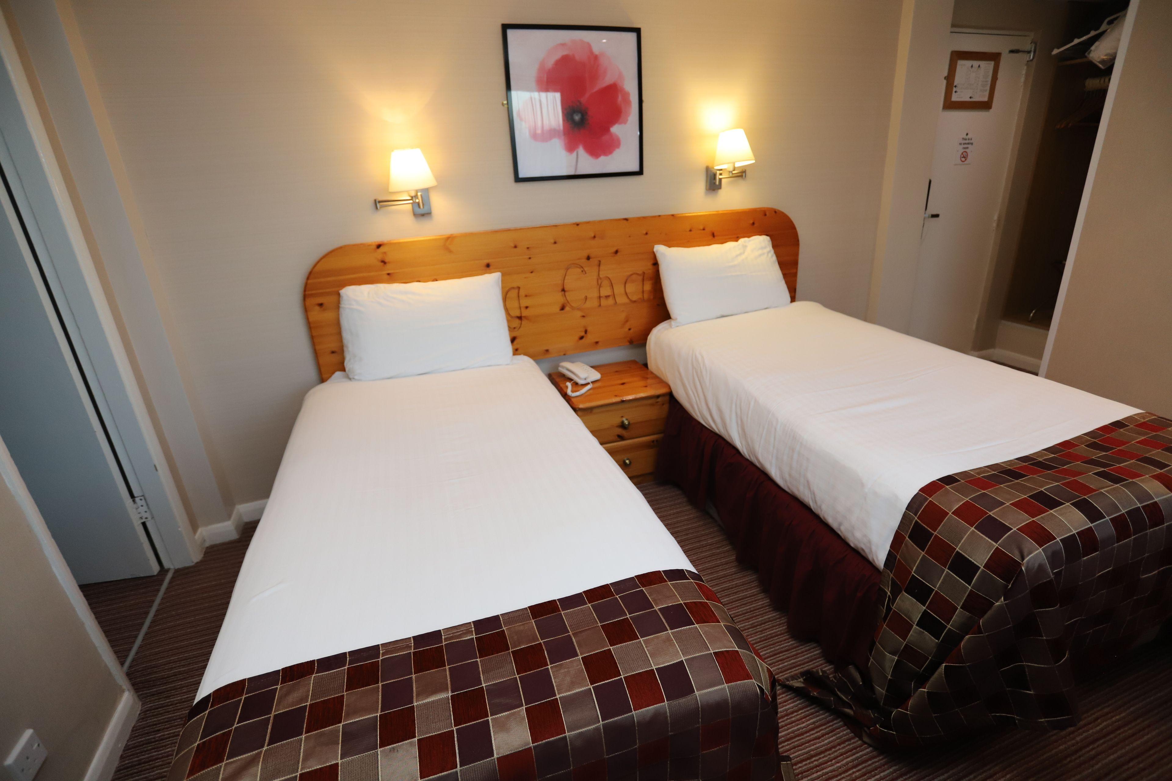 A hotel room with two single beds, a wooden headboard, bedside lamps, a framed artwork, and a door with a sign.