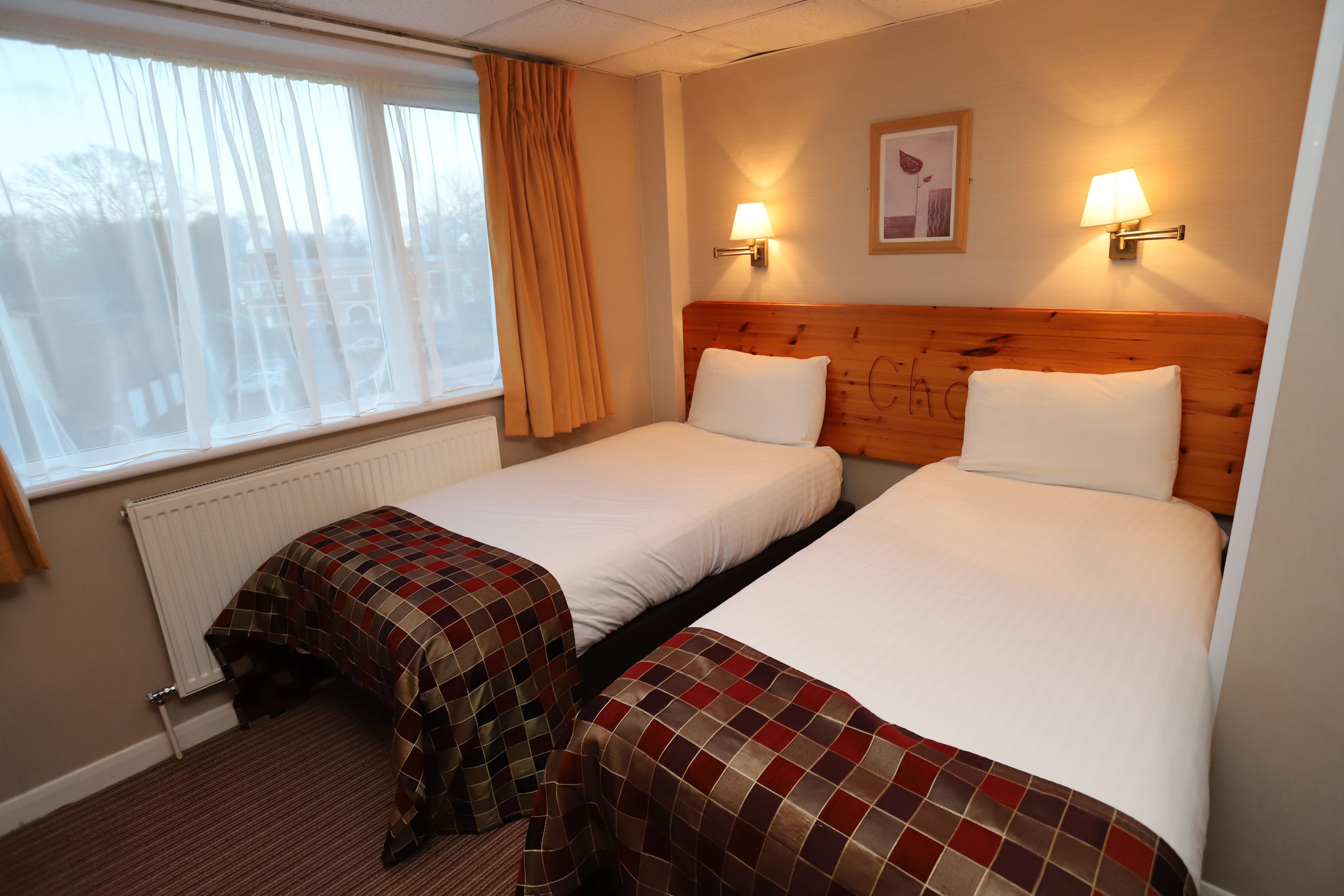 Una habitación de hotel con dos camas, una ventana con cortinas, un radiador y dos lámparas fijadas a la pared.