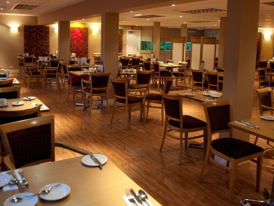 A spacious restaurant with multiple dining tables and chairs, wooden flooring, and decorative wall panels.