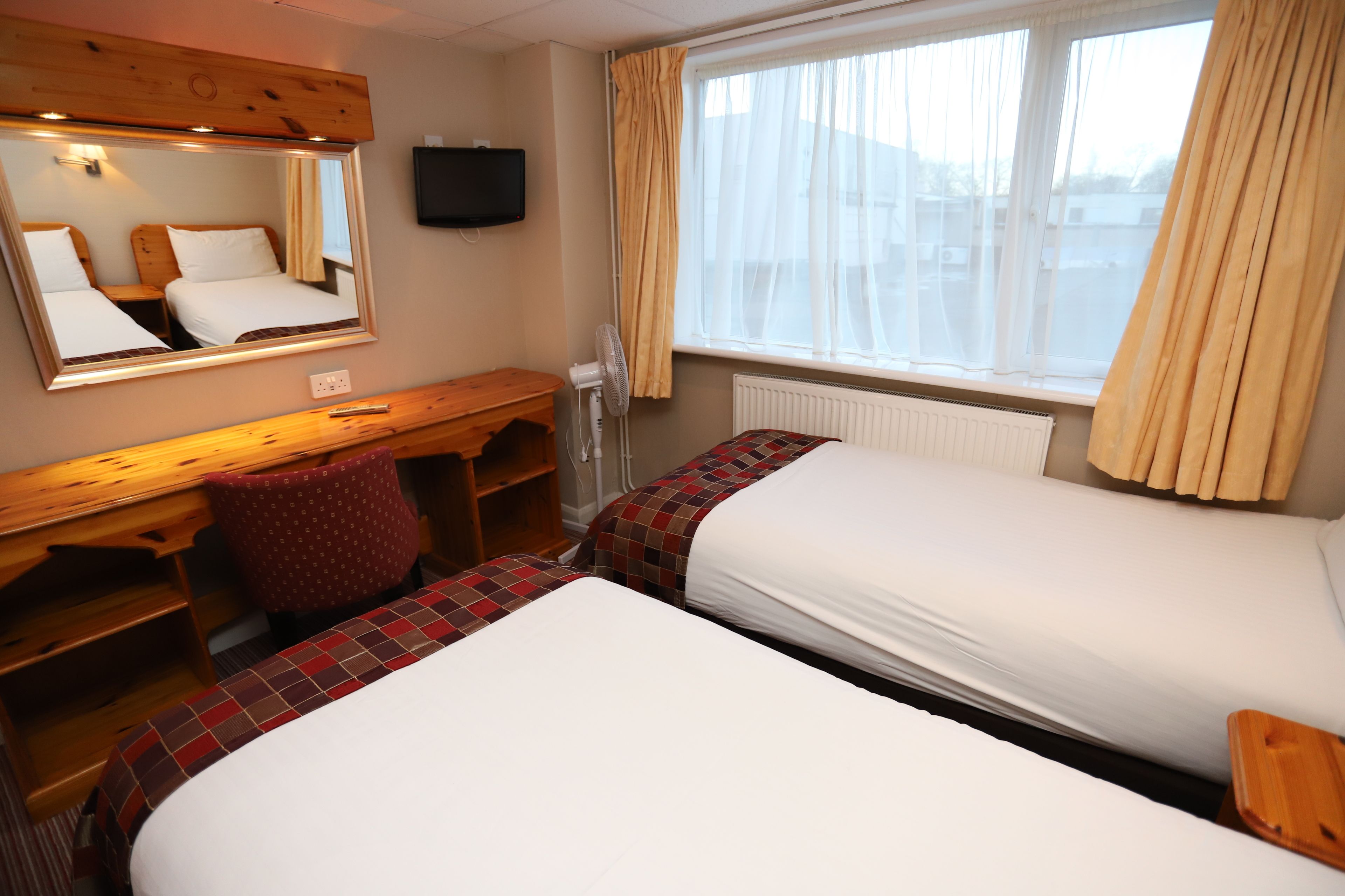 A hotel room with two beds, a wooden desk, a mirror, a TV, and a window with curtains.
