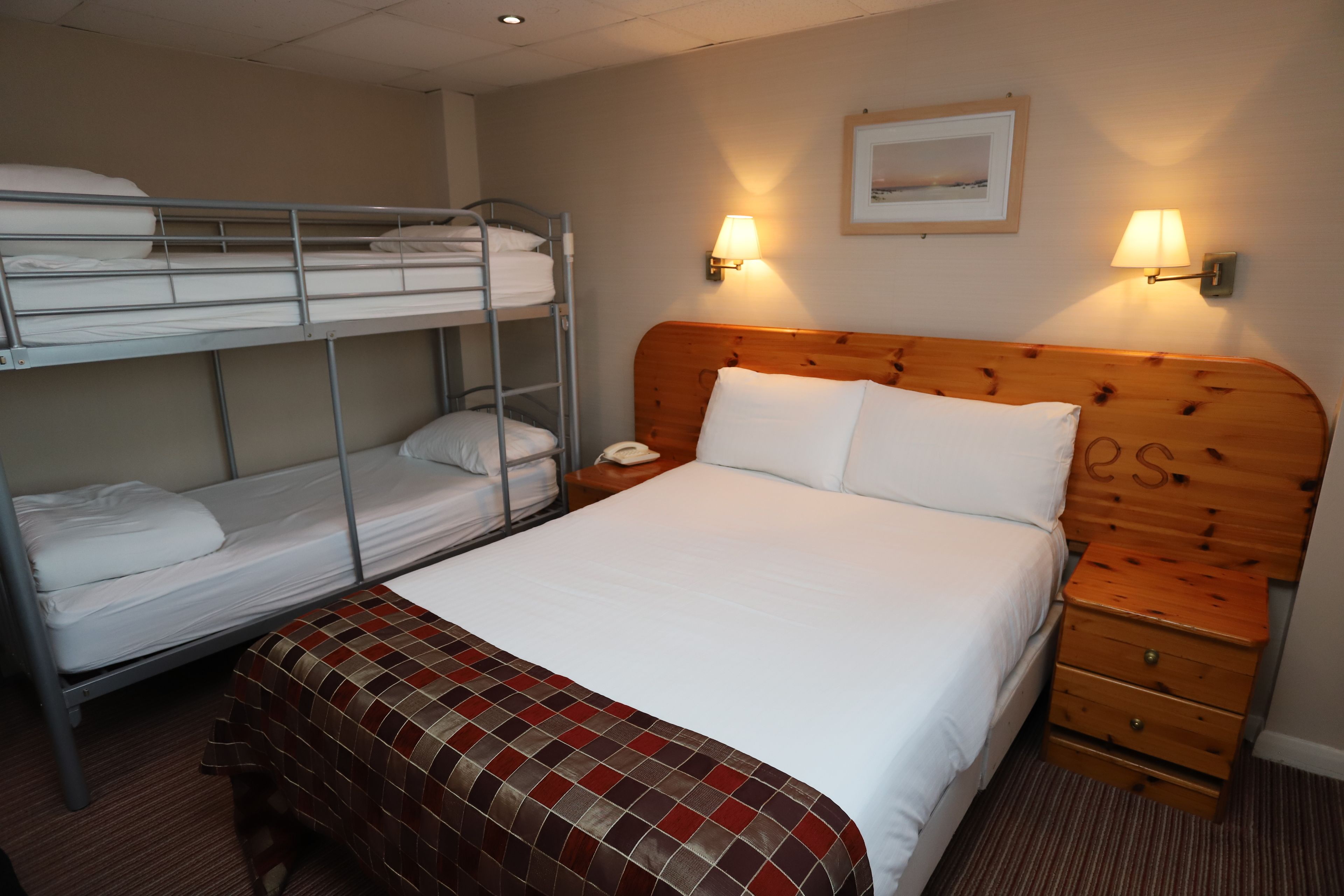 A hotel room with a bunk bed, a single bed, a wooden nightstand, and two wall-mounted lamps.