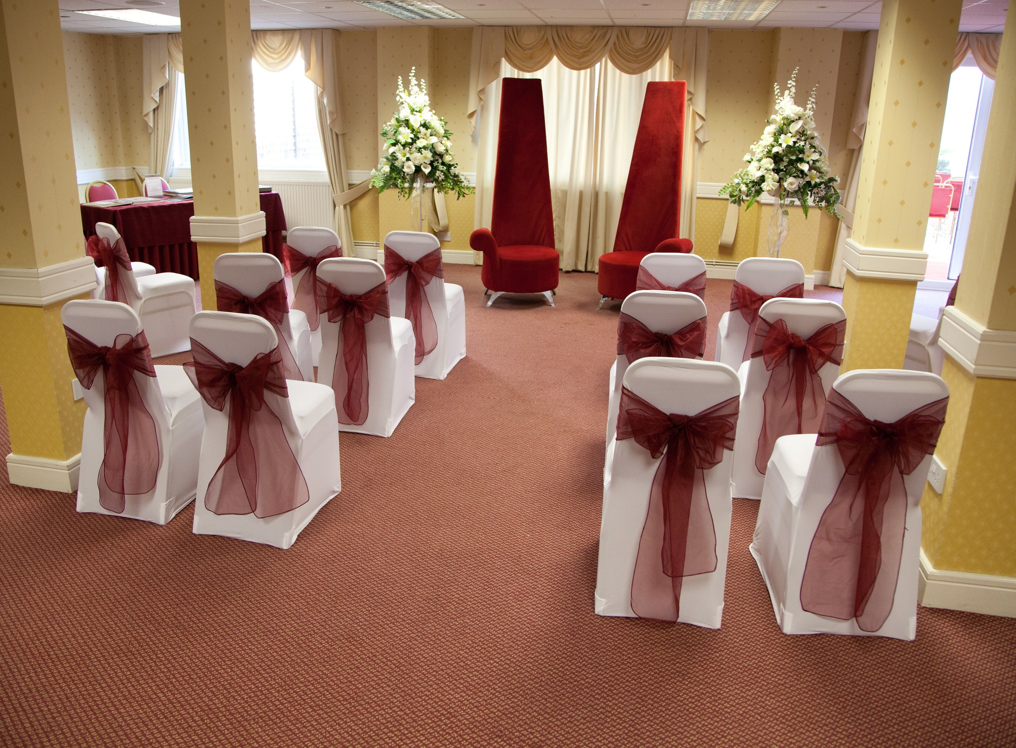 Ein Raum mit Reihen weißer Stühle mit roten Bändern, einem roten Sofa und zwei Blumenarrangements.