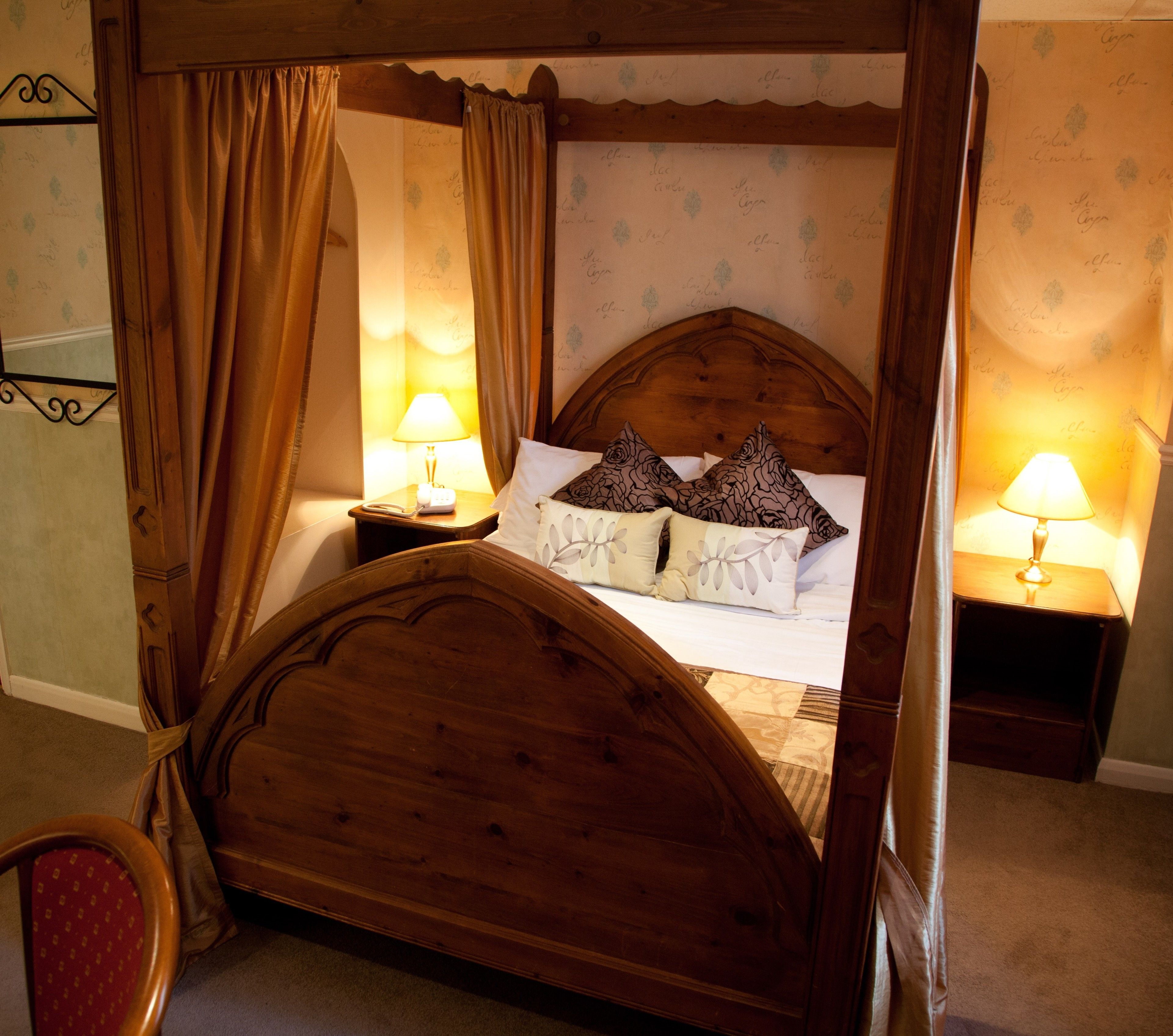 A four-poster bed with a wooden headboard, two bedside tables with lamps, and a floral-patterned wall.