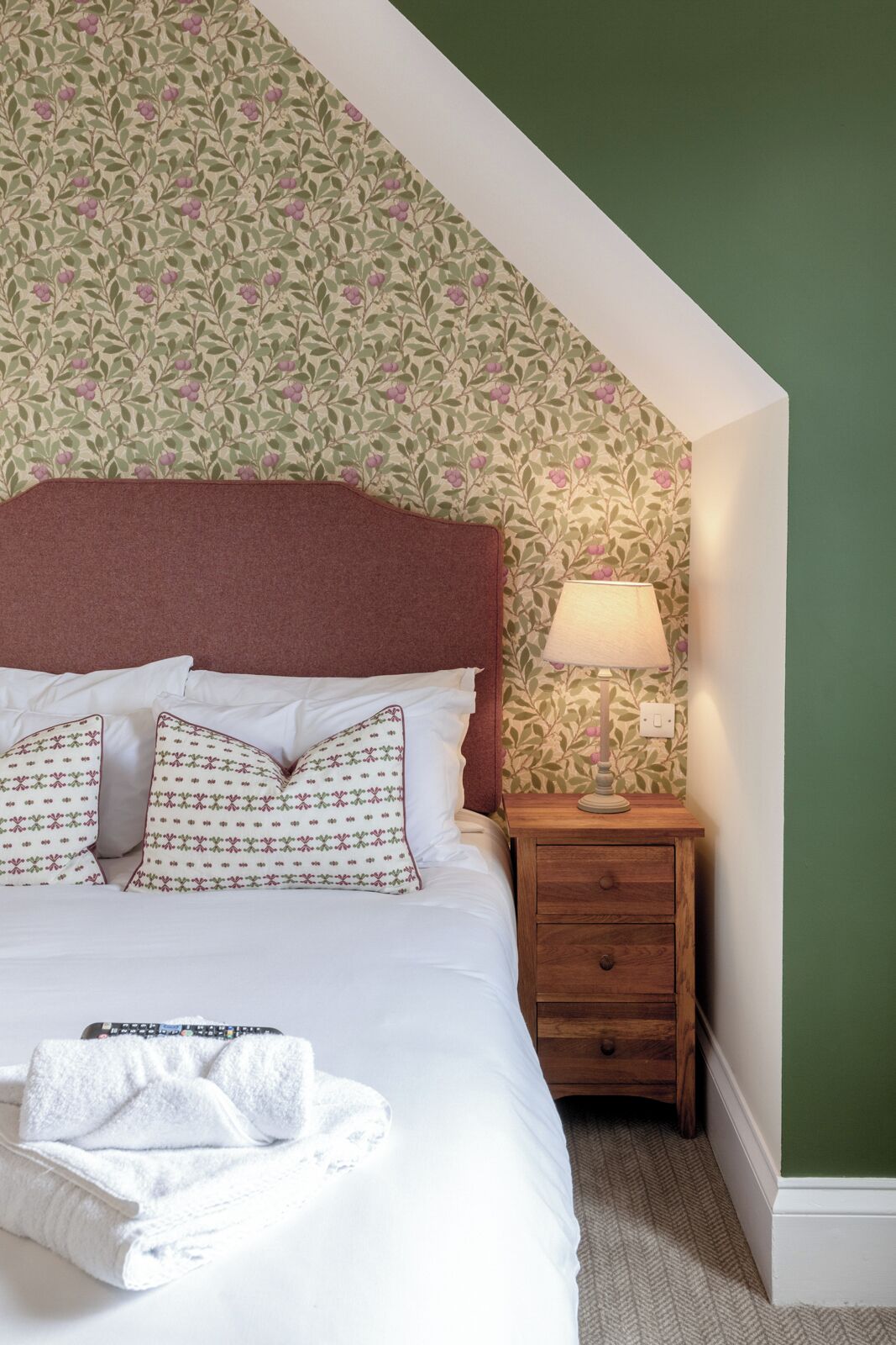 Un lit bien fait, agrémenté d’oreillers à motifs floraux et recouvert d’une couverture blanche, une table de chevet en bois avec une lampe, et un mur vert orné d’un papier peint à motifs floraux.