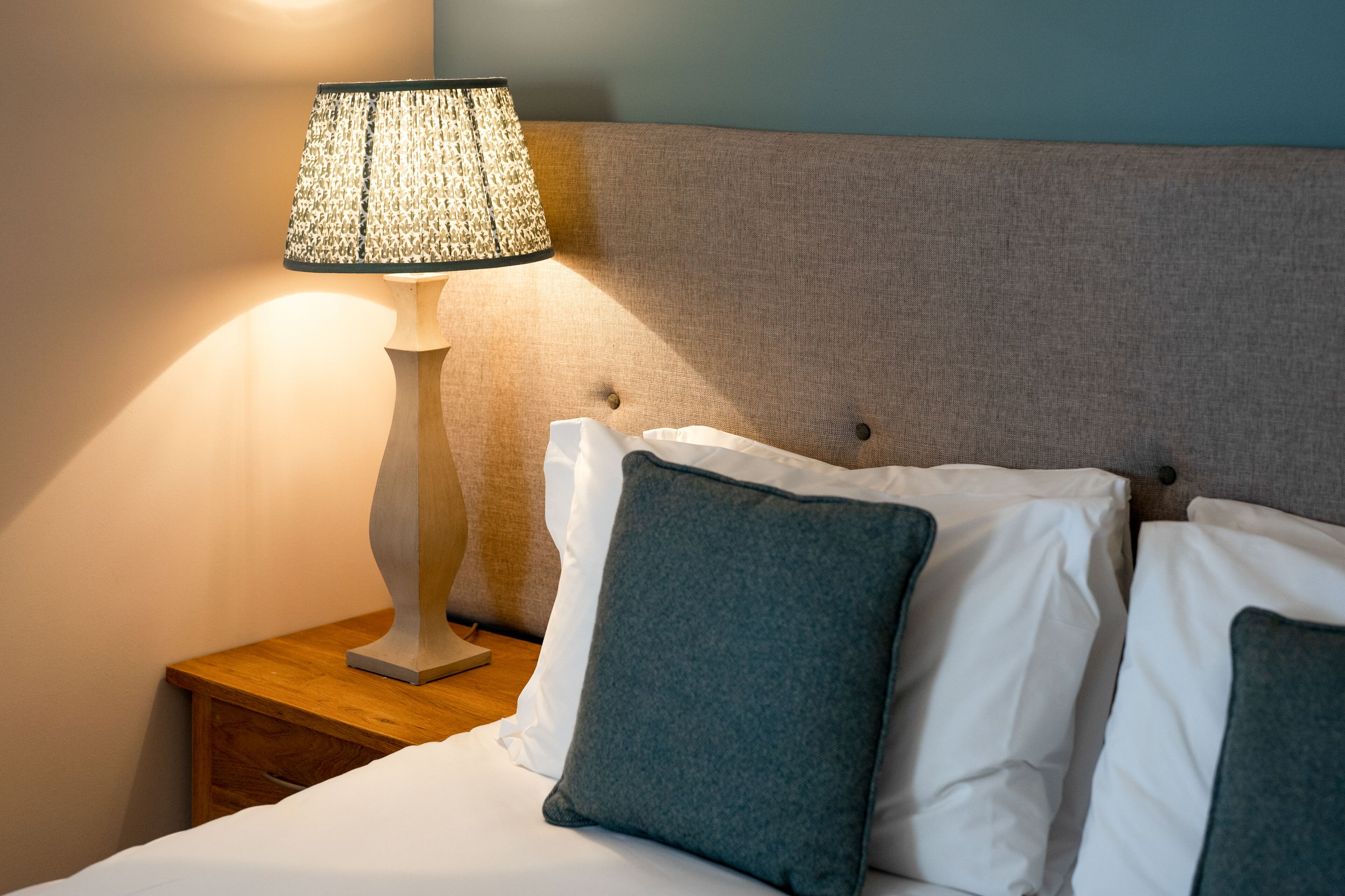 Una cama bien hecha con almohadas blancas y verde oscuro, una lámpara con textura sobre una mesita de noche de madera, y un cabecero con un panel de tela.