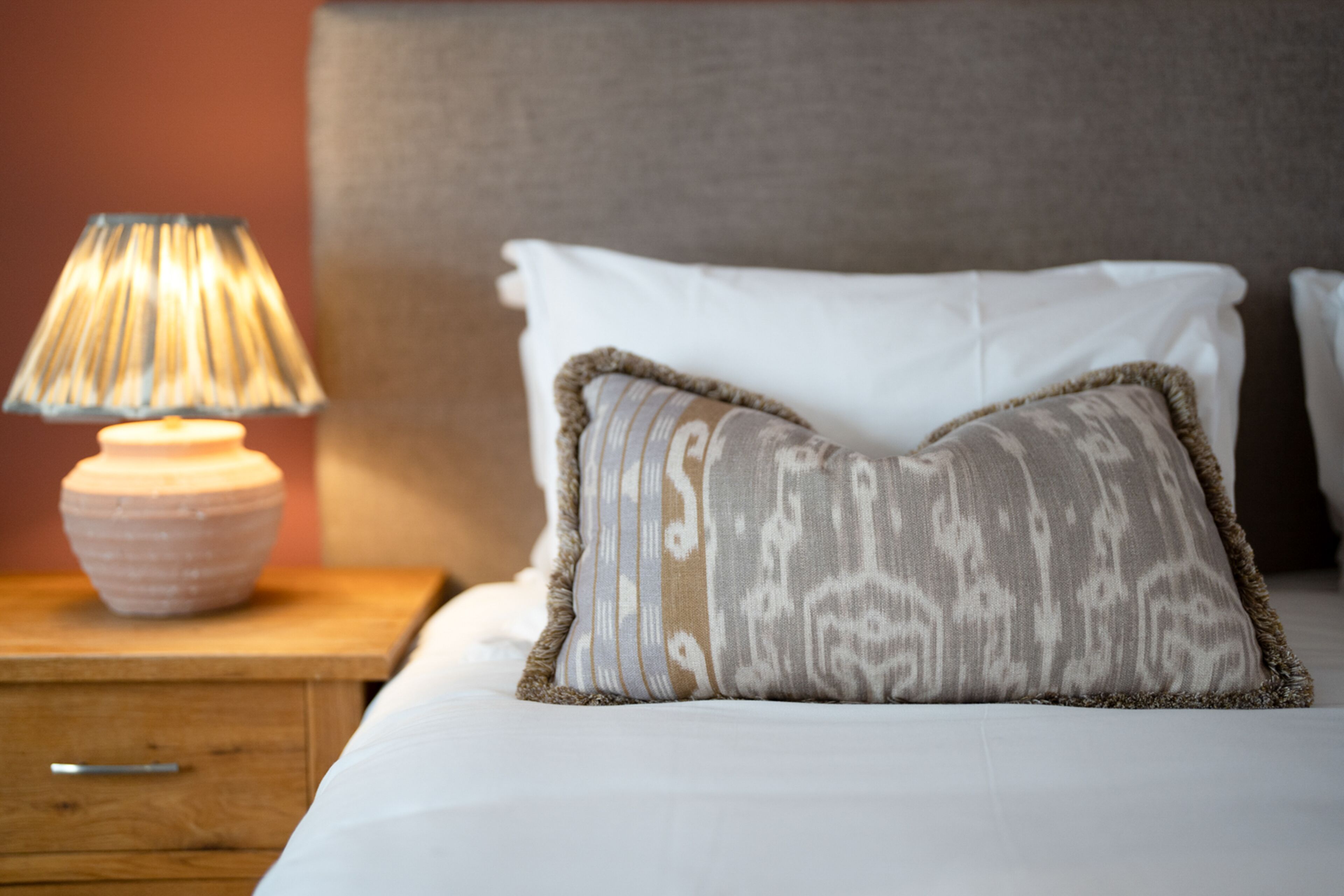 Una cama bien hecha con un cojín estampado y una lámpara de noche sobre una mesita de madera.
