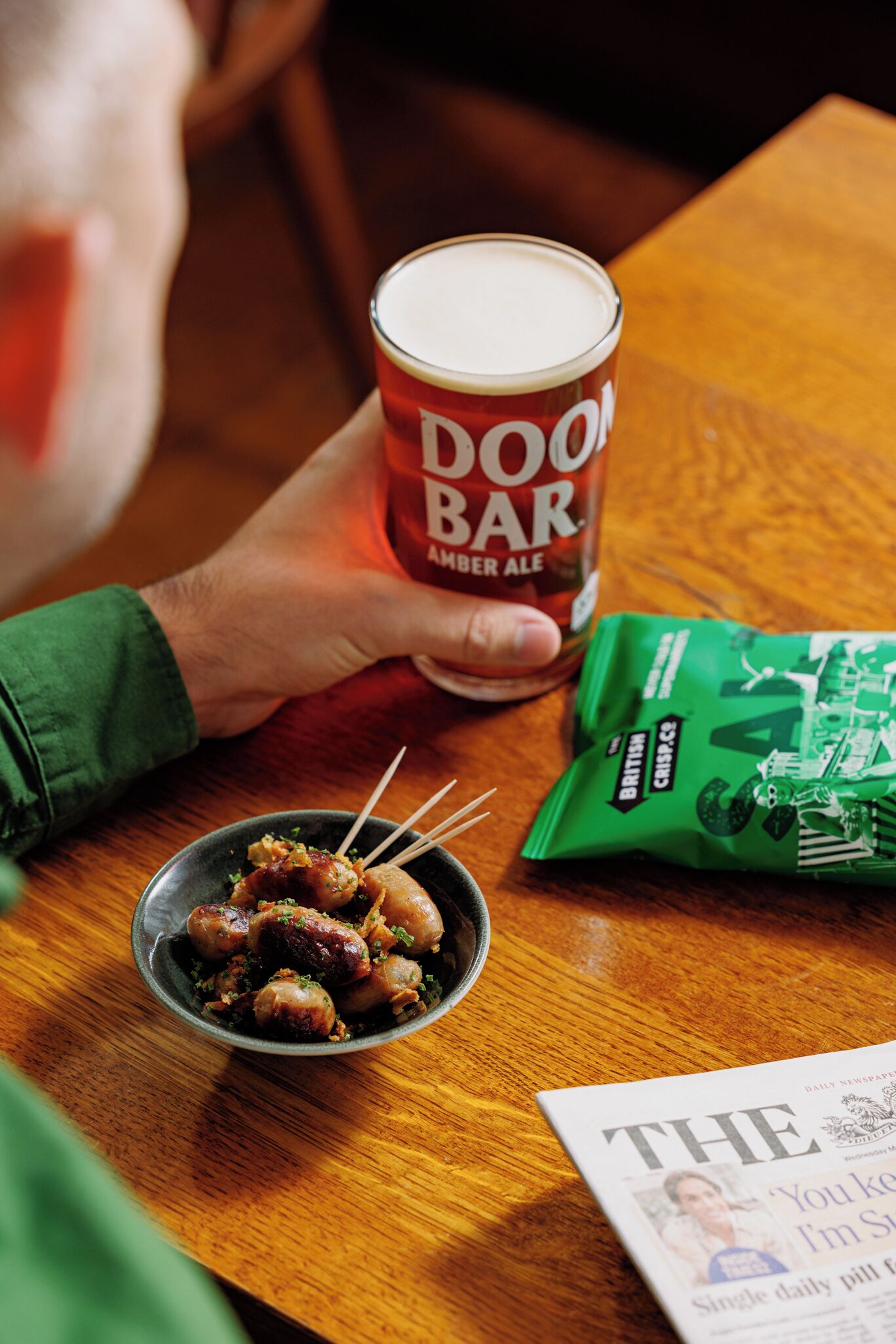 A hand holding a can of Doorn Bar Amber Ale, a bowl of skewered food, and a packet of crisps on a wooden table.