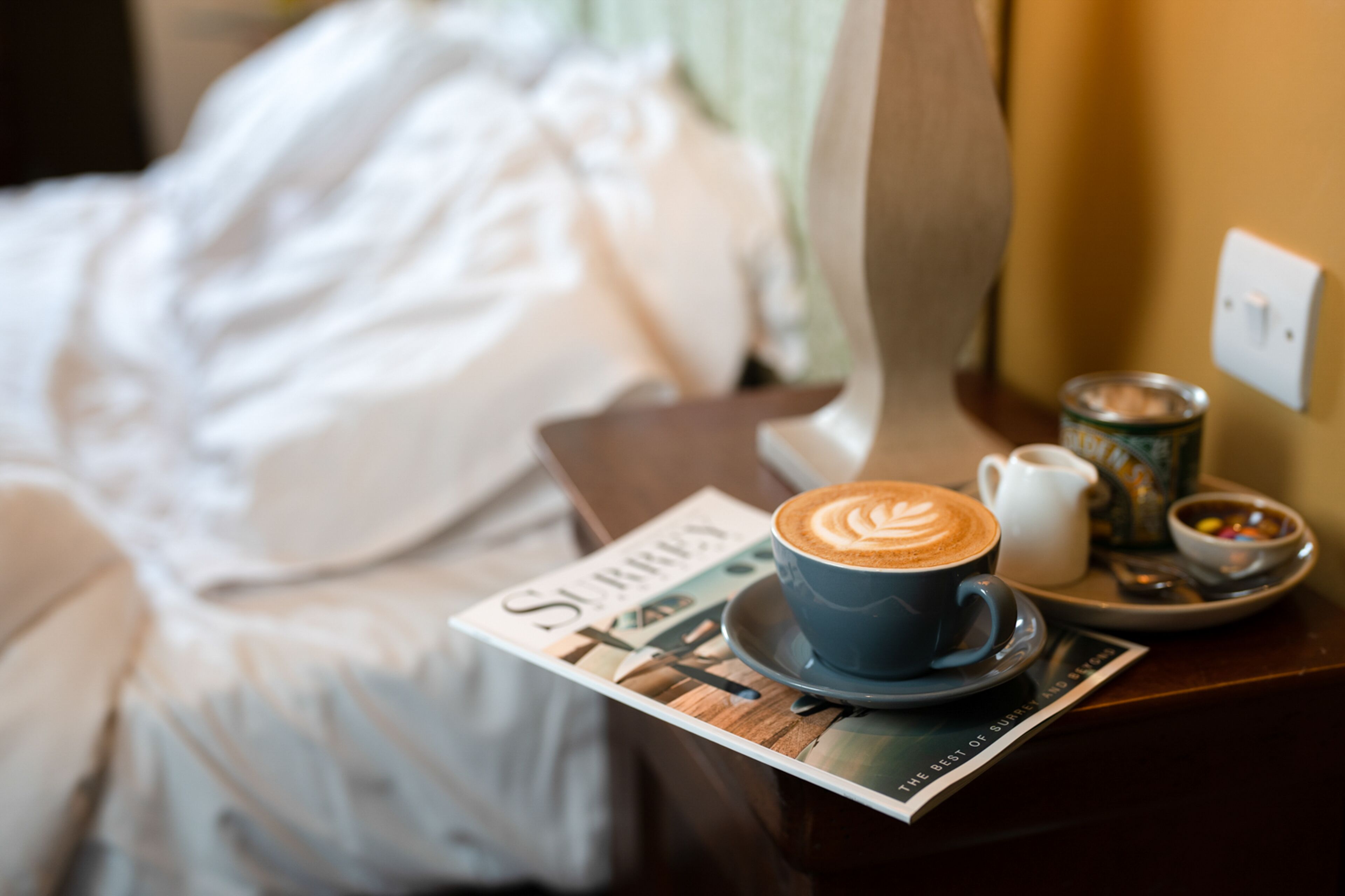 A bed with white linens, a coffee cup with latte art, a newspaper, and a tray with a cup of coffee and a small bowl of candies.