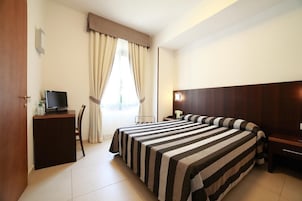 A hotel room with a large bed, a desk with a computer, a chair, and a window with curtains.