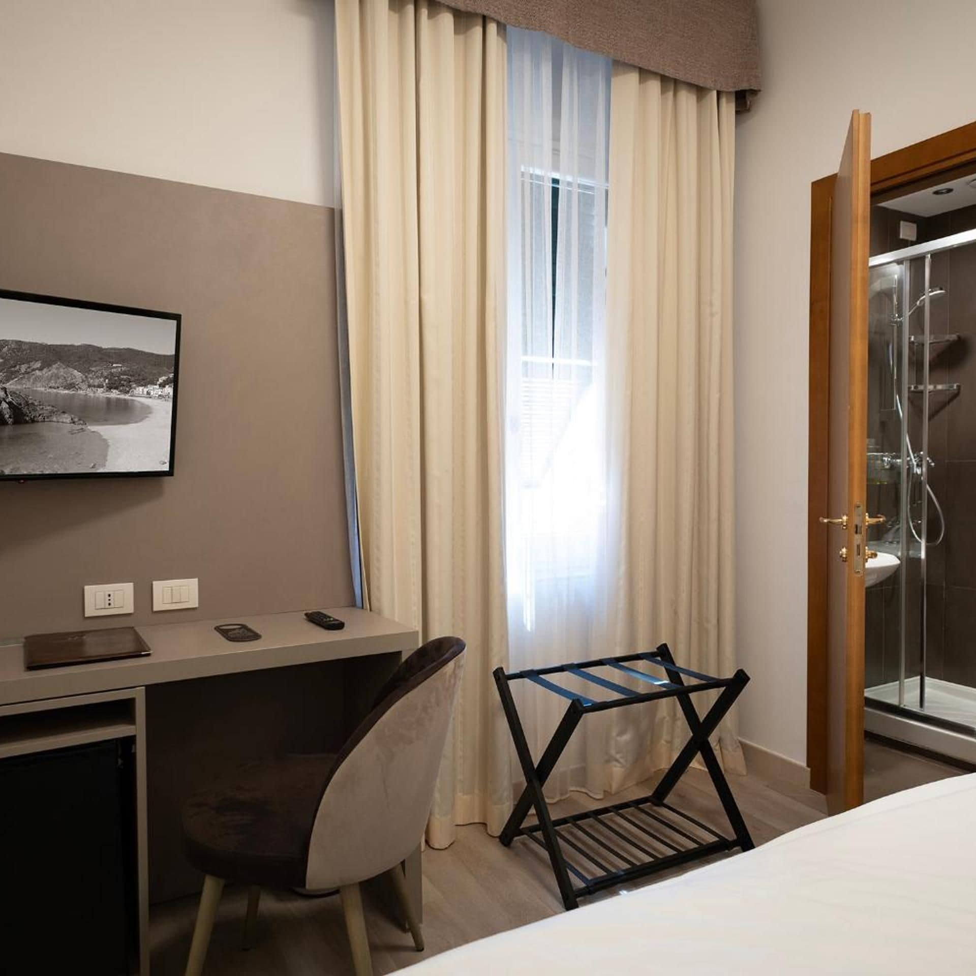 A hotel room with a bed, a desk with a chair, a television, a window with curtains, and a shower area.