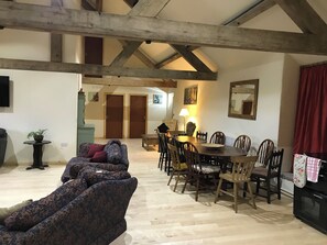 A spacious living room with a high ceiling and exposed beams, a dining area with wooden chairs and a table, and a cozy seating area with patterned sofas. - Witherslack Hall Farm (Grange-over-Sands)