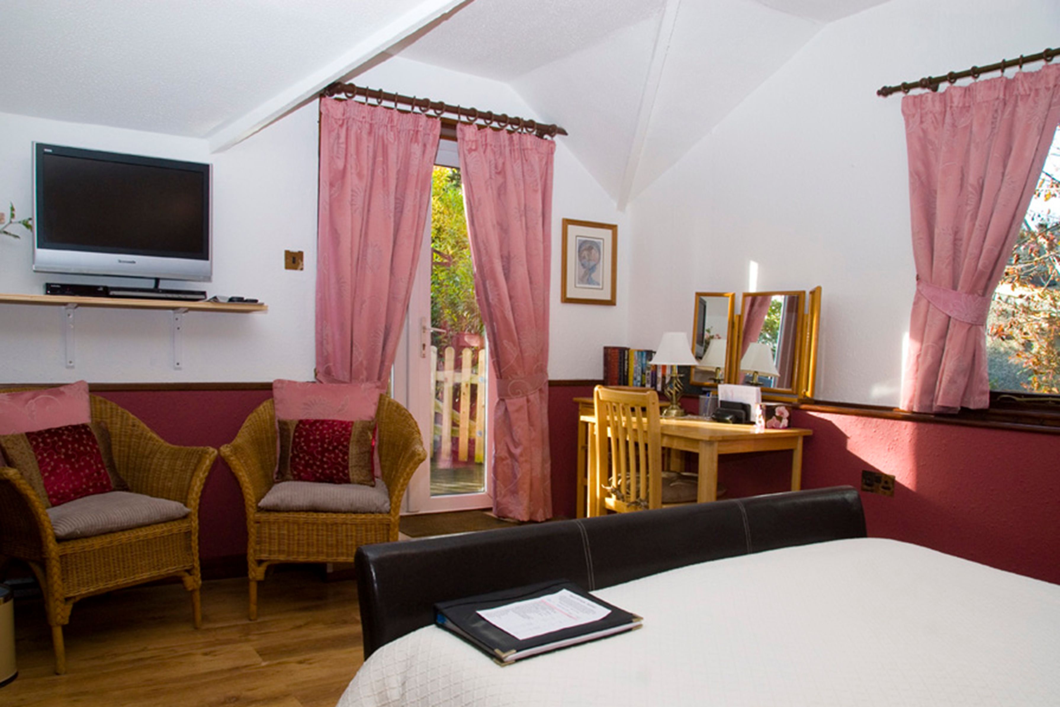 A bedroom with a bed, a television on a shelf, wicker chairs, a desk with a mirror, and pink curtains.