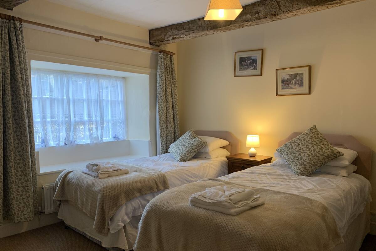 Ein Hotelzimmer mit zwei Betten, einem Fenster mit transparenten Vorhängen, einer Nachttischlampe und zwei gerahmten Bildern an der Wand.