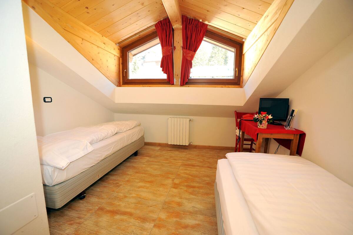 A small, single-room apartment with a sloped ceiling, a window with red curtains, a radiator, a desk with a computer, and two beds.