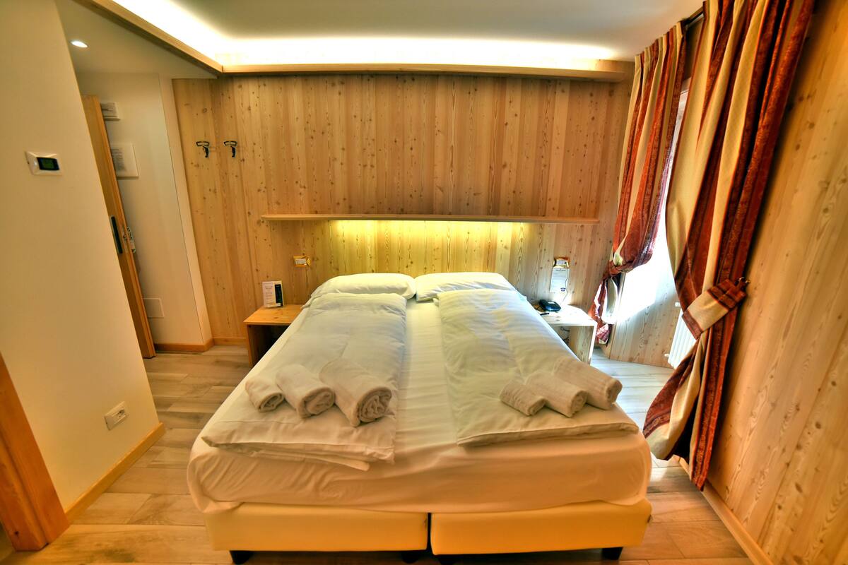 A double bed with white linens and towels, wooden paneling on the walls, a bedside table with a lamp, and a window with curtains.