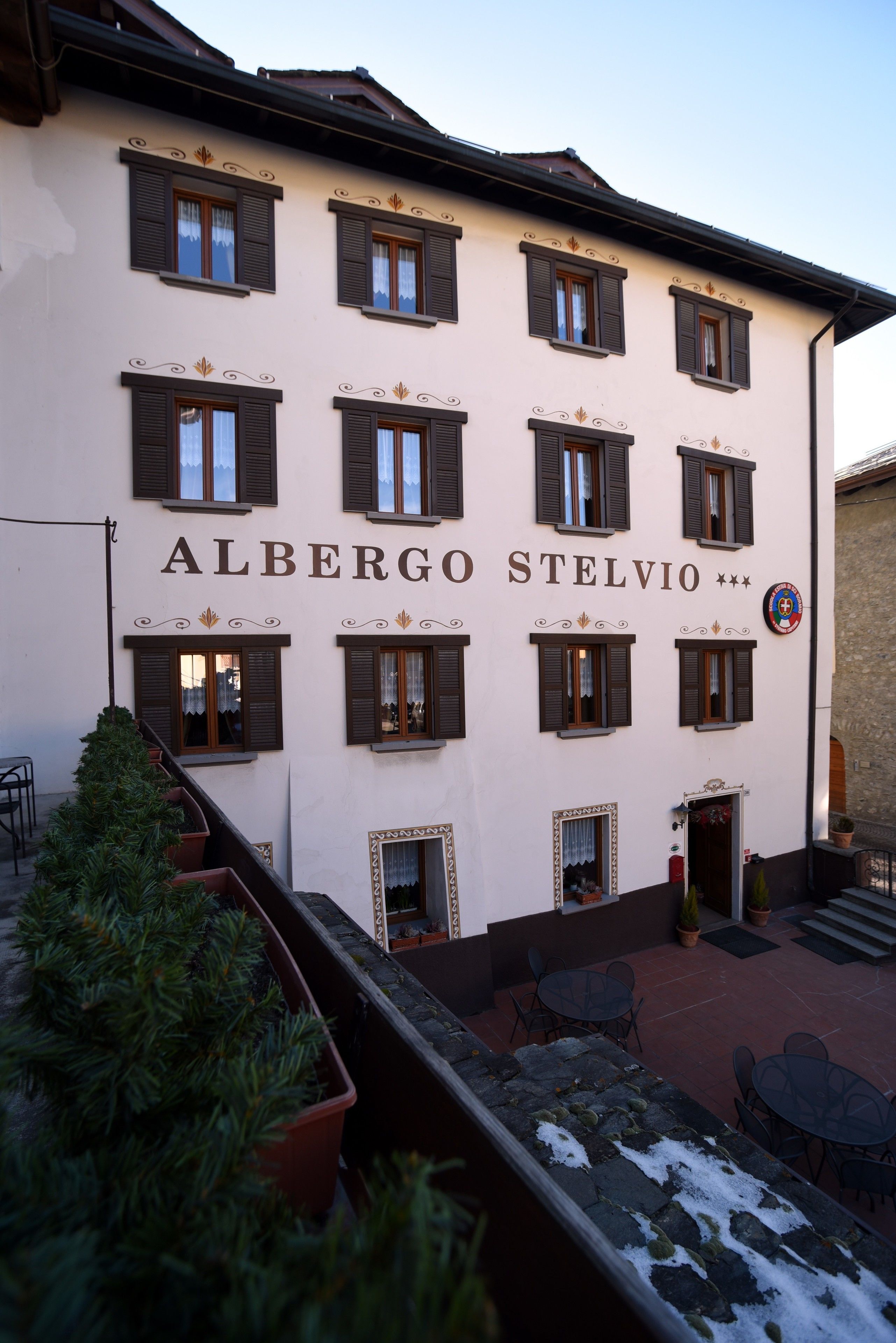 A building with the name "ALBERGO STELVIO" on its facade, featuring multiple windows with shutters, a stone staircase, and outdoor seating.