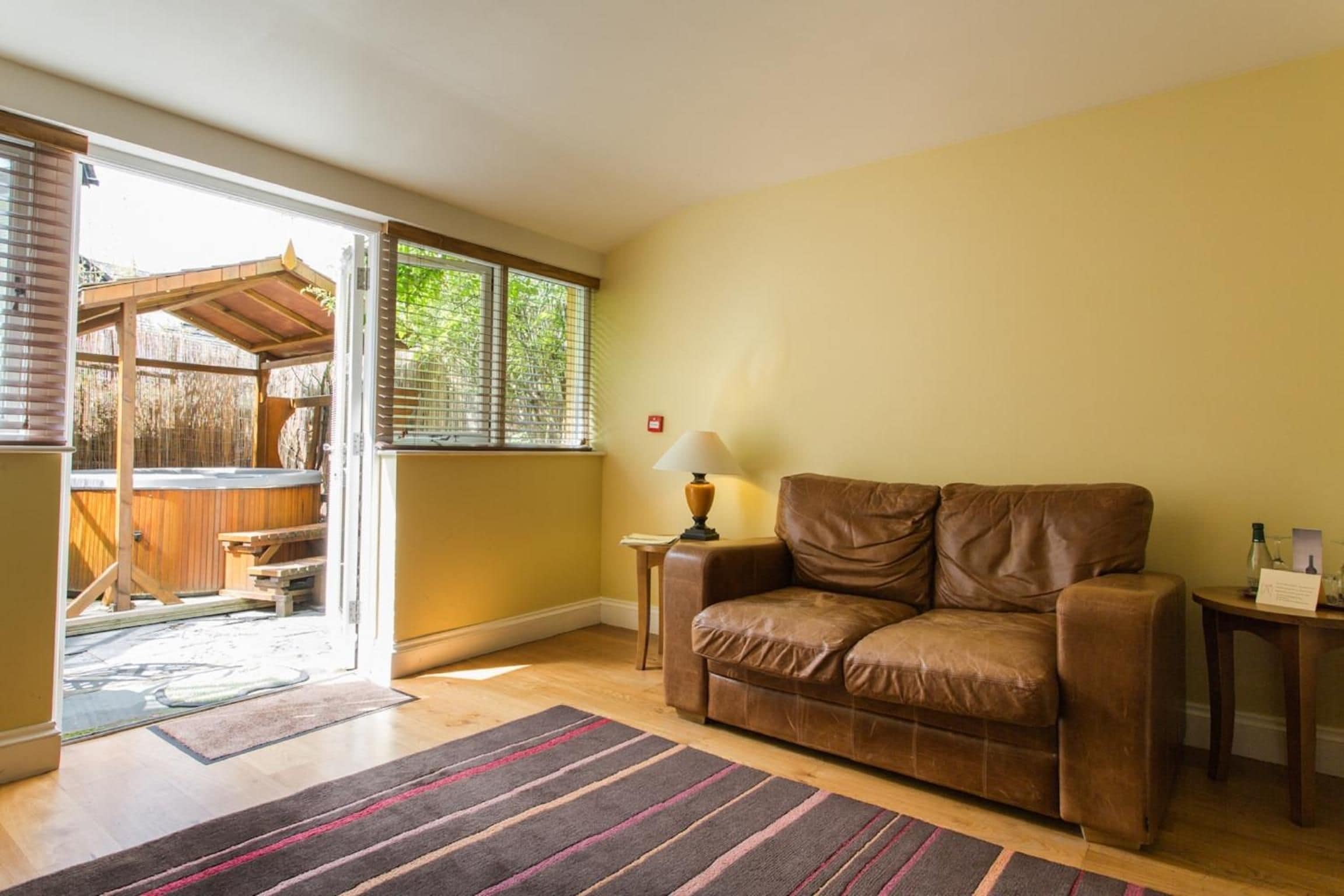A living room with a brown leather sofa, a lamp on a side table, and a view of an outdoor area with a wooden structure.