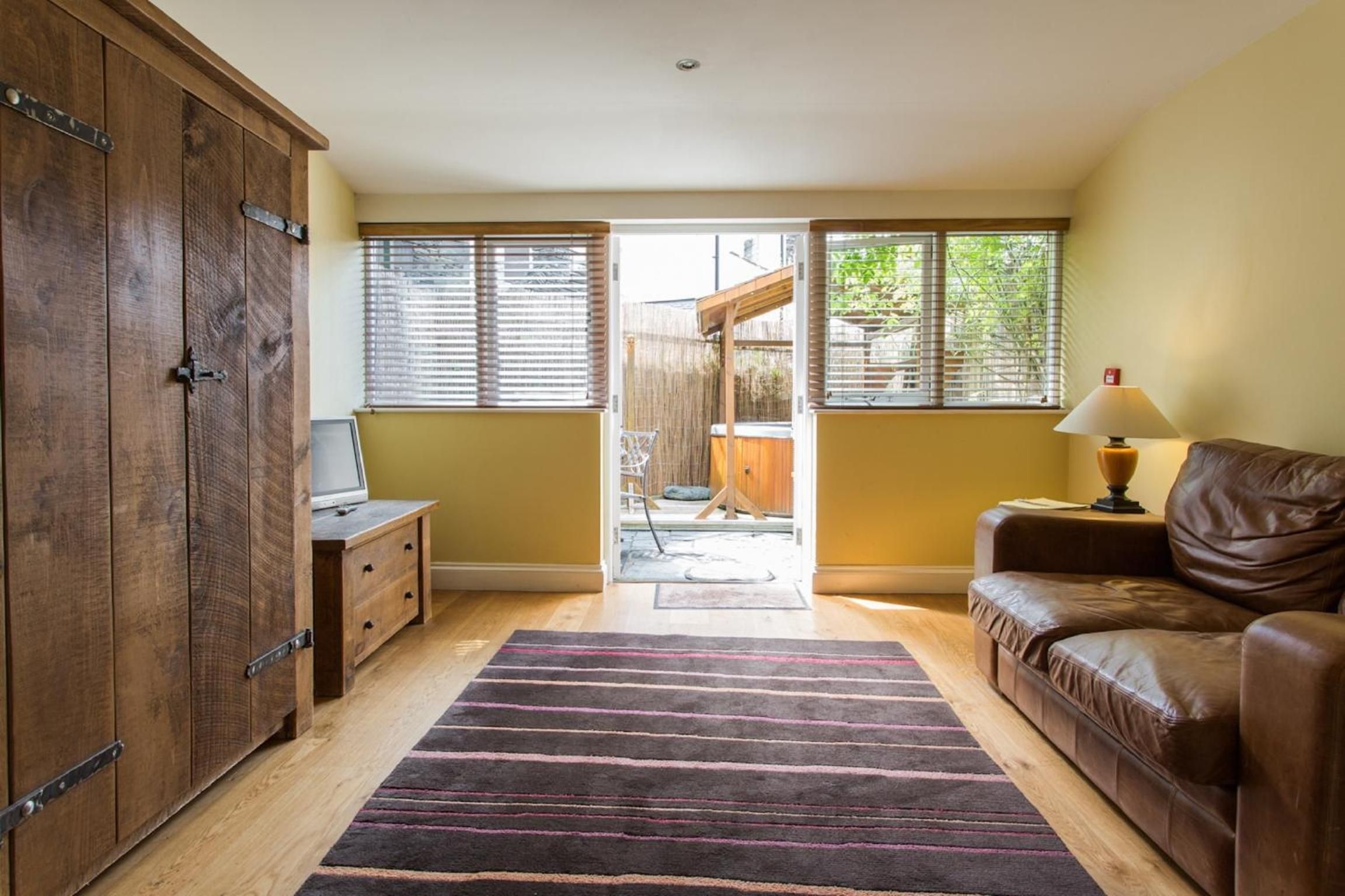 A room with a wooden wardrobe, a leather sofa, a small table with a lamp, and a striped rug.