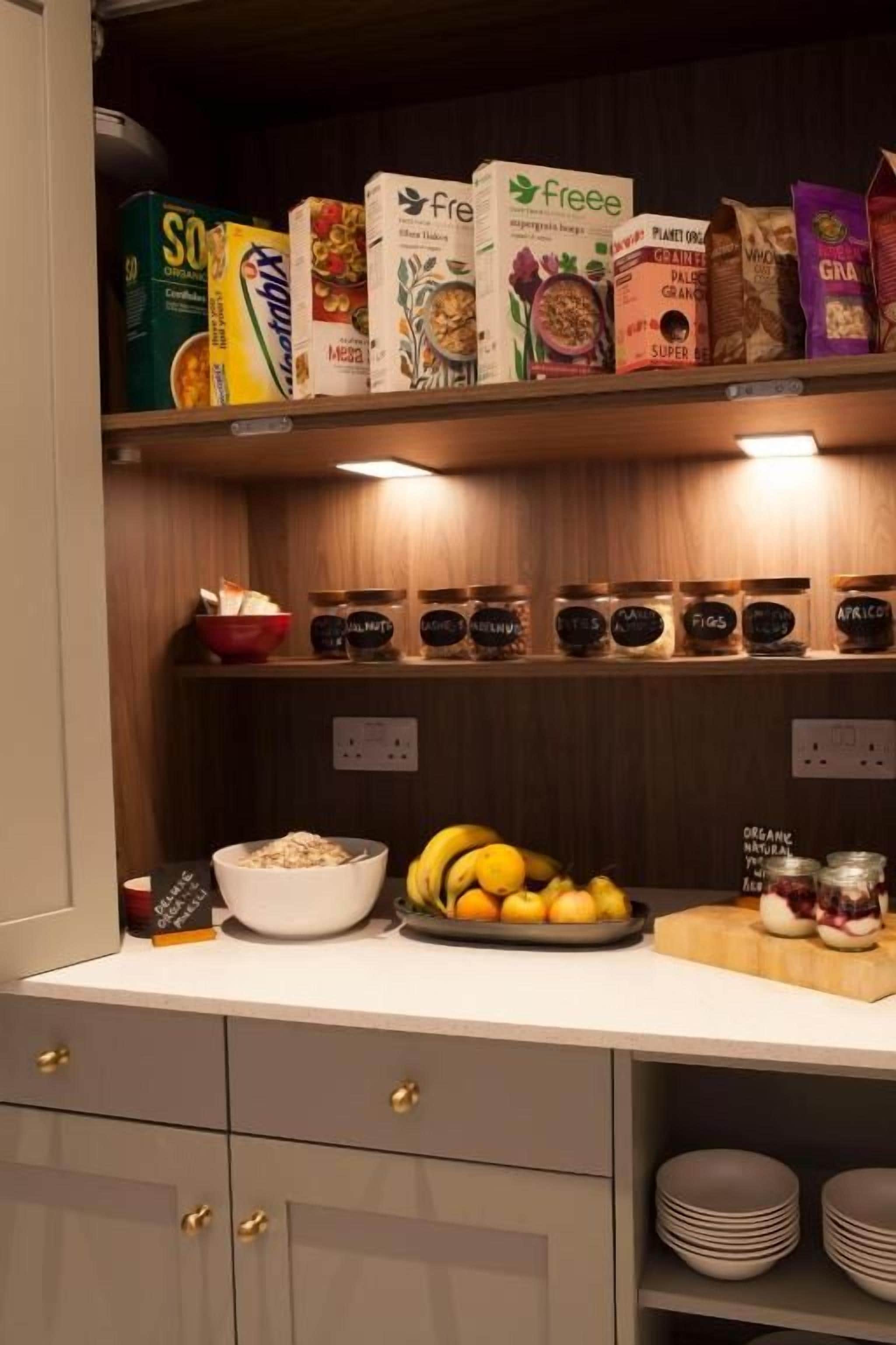 A kitchen with shelves stocked with food items, a bowl of fruit, and a plate of sliced fruit.