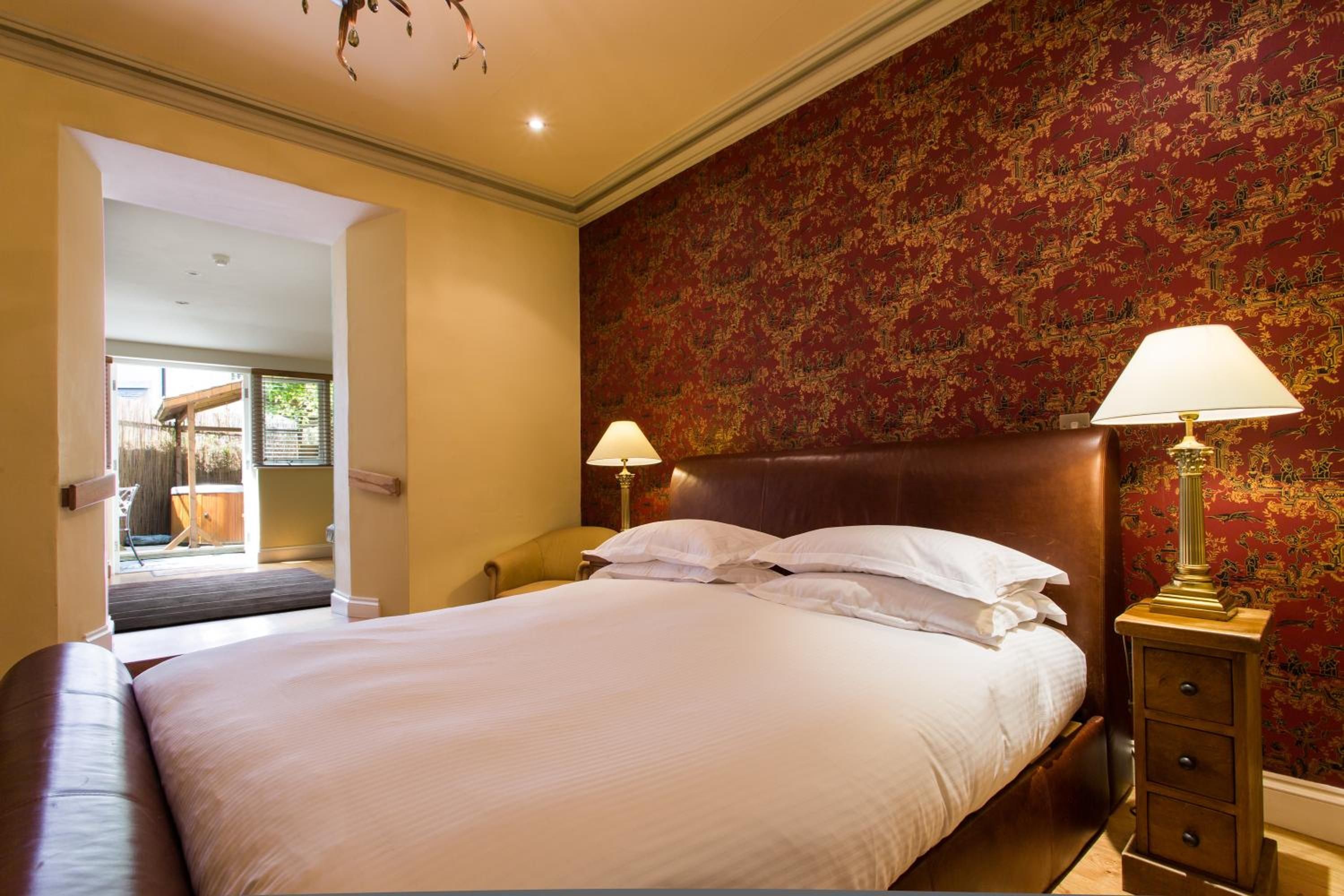 A hotel room with a large bed, two bedside lamps, a wooden nightstand, and a red patterned wallpaper.