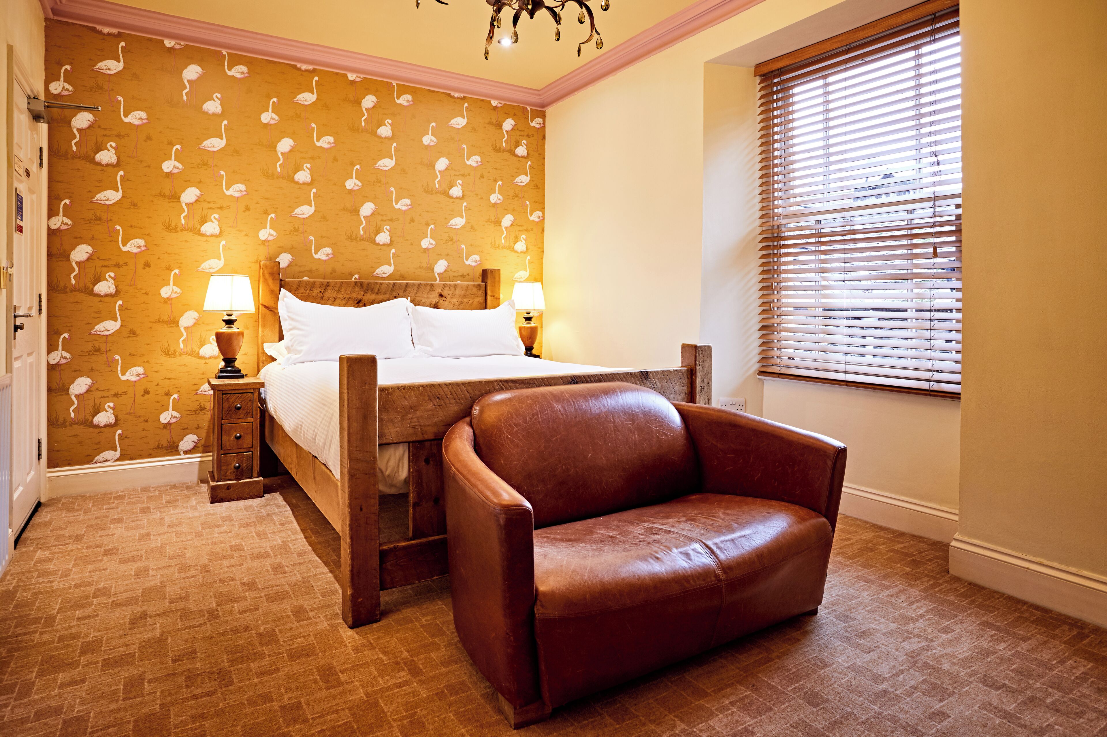 A bedroom with a bed, a wooden nightstand, a brown leather armchair, a patterned wallpaper, and a window with blinds.