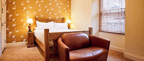 A bedroom with a bed, a wooden nightstand, a brown leather armchair, a patterned wallpaper, and a window with blinds.
