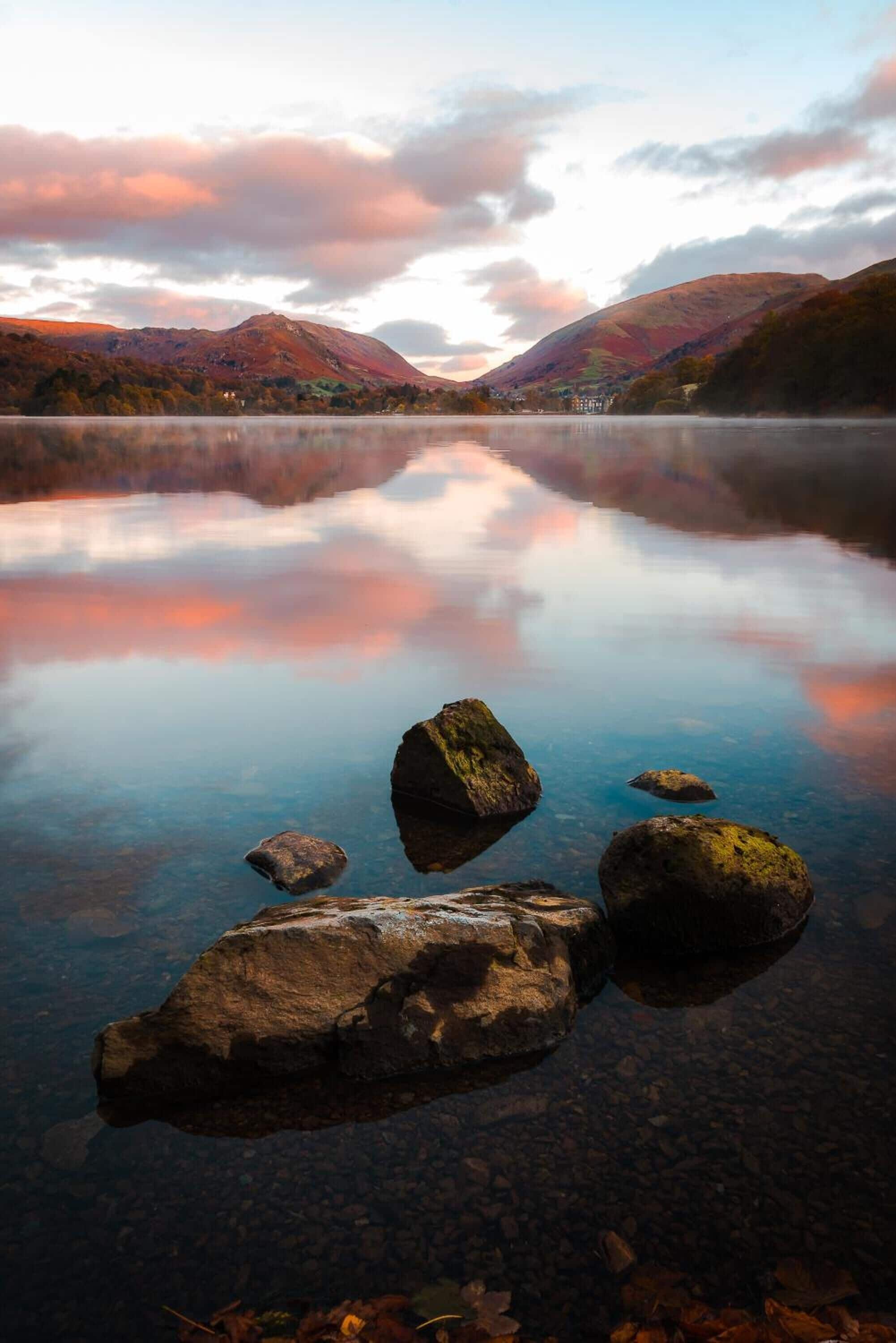 A tranquil lake with clear reflections, surrounded by mountains and a house in the distance.