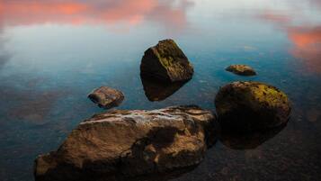 A tranquil lake with clear reflections, surrounded by mountains and a house in the distance.