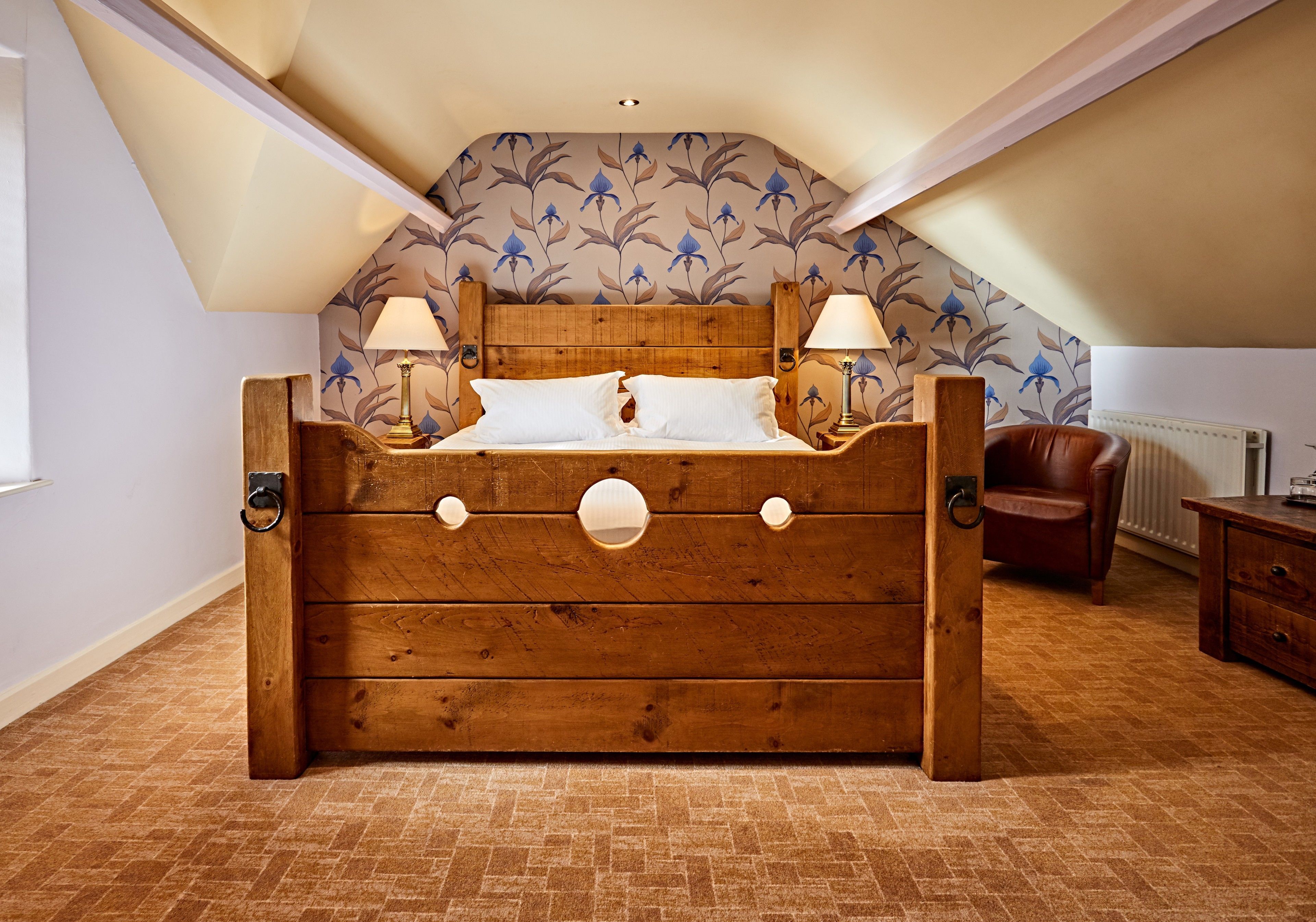A wooden bed with a sloped ceiling and floral wallpaper, a brown leather chair, and a wooden nightstand.