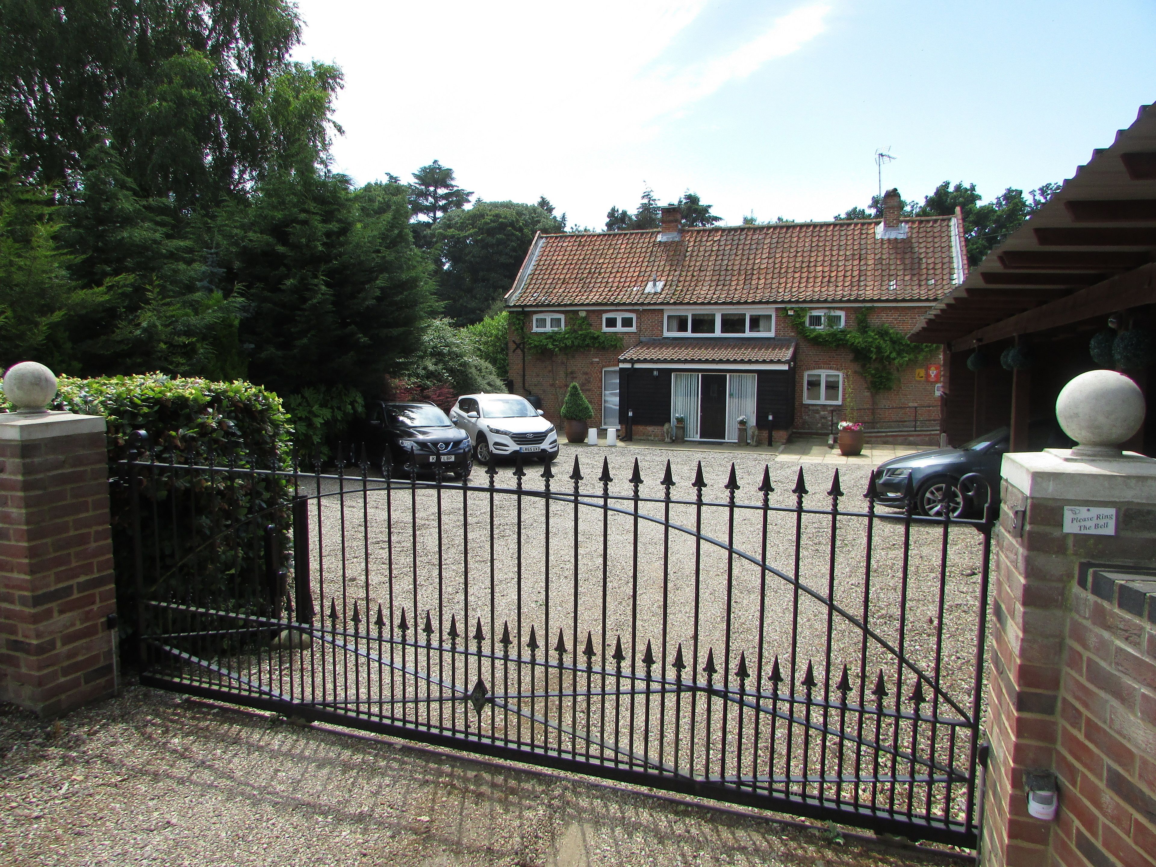 A gated driveway leading to a brick house with a tiled roof, two parked cars, and a sign that says "Please Ring The Bell".