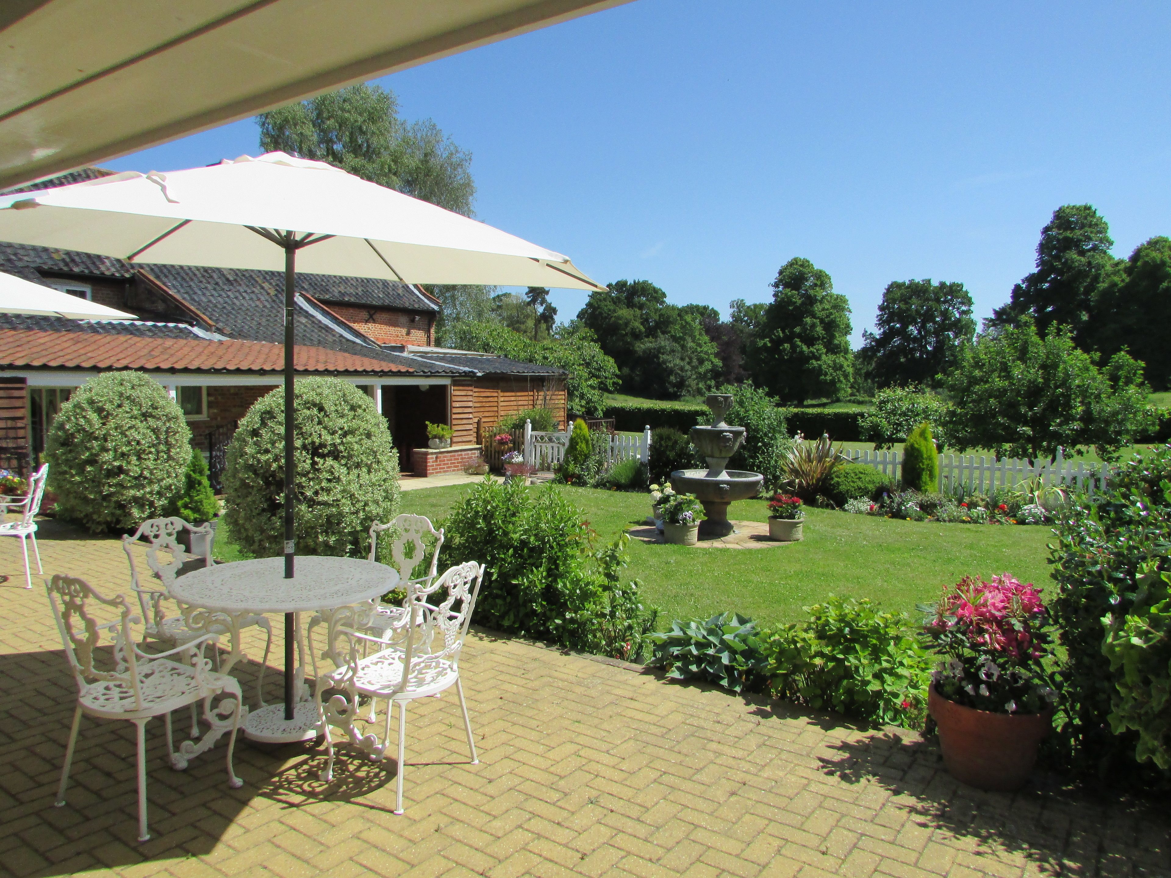 En terrasse med et bord og stole, en have med en springvand, og et hus i baggrunden.