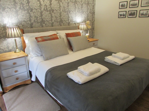 A neatly made bed with a grey bedspread, white pillows, and two folded white towels. There are two bedside tables with lamps and a small nightstand with a lamp. The room has a patterned wallpaper and a framed picture on the wall. - Decoy Barn (Great Yarmouth)