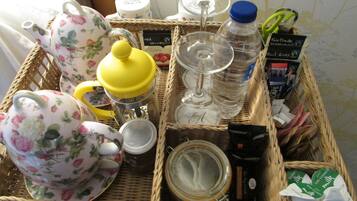 A wicker basket containing various items including a floral teapot, a yellow container, a glass, a jar, and some packaged food items.