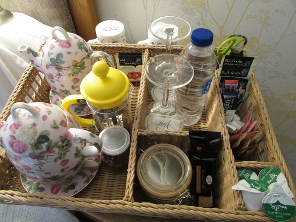 A wicker basket containing various items including a floral teapot, a yellow container, a glass, a jar, and some packaged food items. - Decoy Barn (Great Yarmouth)