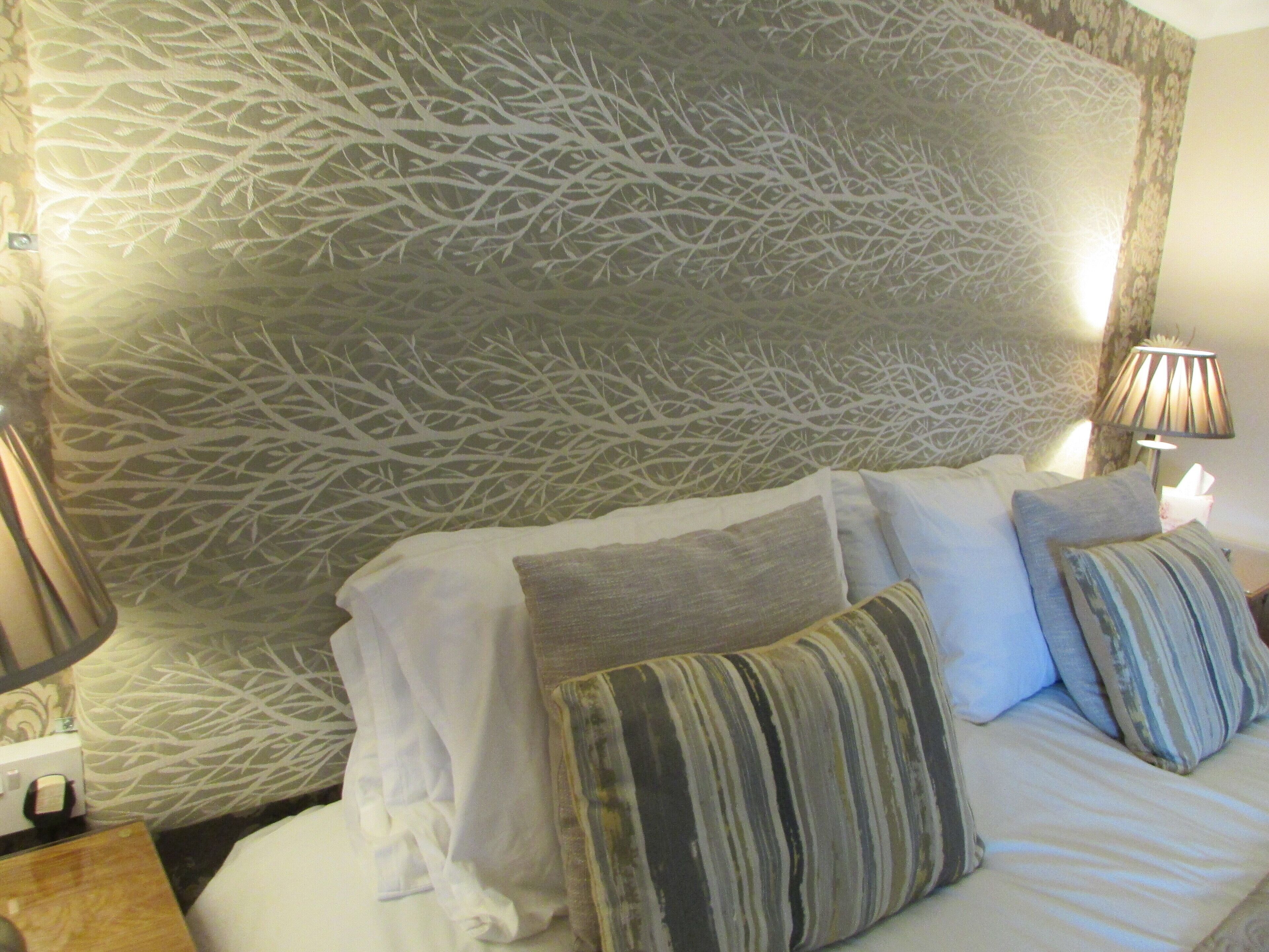 A hotel room with a patterned wall, a bed with multiple pillows, a bedside lamp, and a nightstand with a tissue box.
