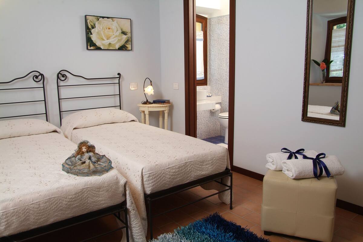 A bedroom with two beds, a small bedside table with a lamp, a mirror, and a bathroom visible through an open door.