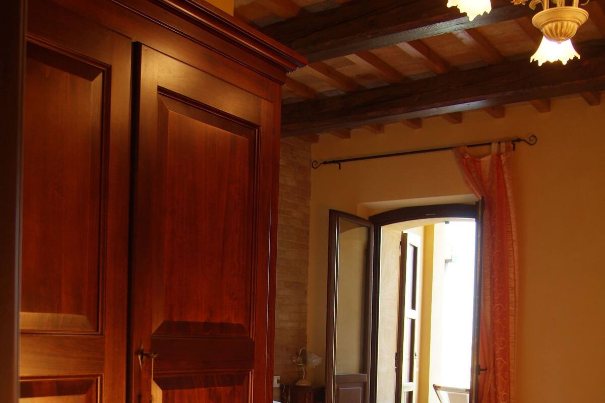 A bedroom with a wooden wardrobe, a bed with a white bedspread, and a view of the outside through an open door.