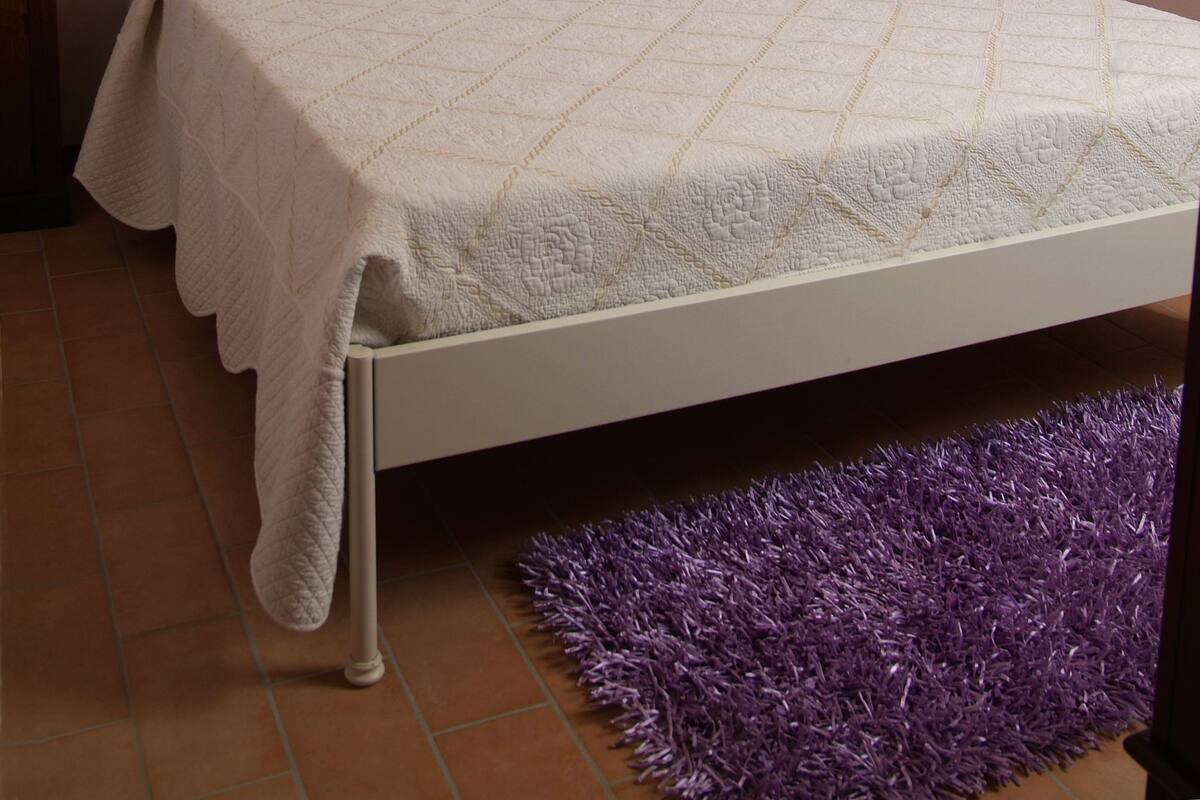 A bedroom with a bed, a purple rug, a wall with a decorative item, and a shelf with a plant.