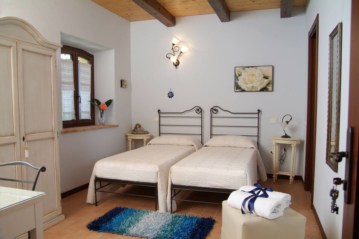 A bedroom with two single beds, a bedside table with a lamp, a window with curtains, a painting on the wall, and a door with a key in the lock.