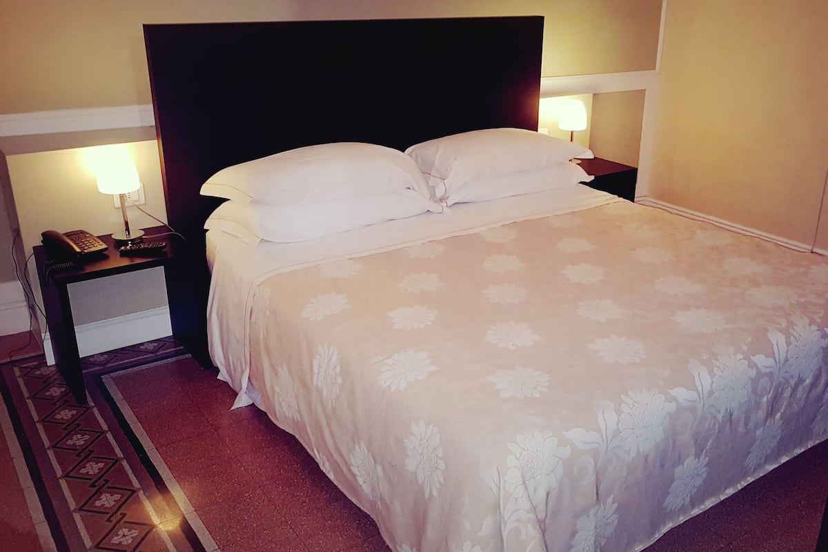Een net opgemaakt bed met een bloemenbedekking, een bedlampje en een telefoon op het nachtkastje.