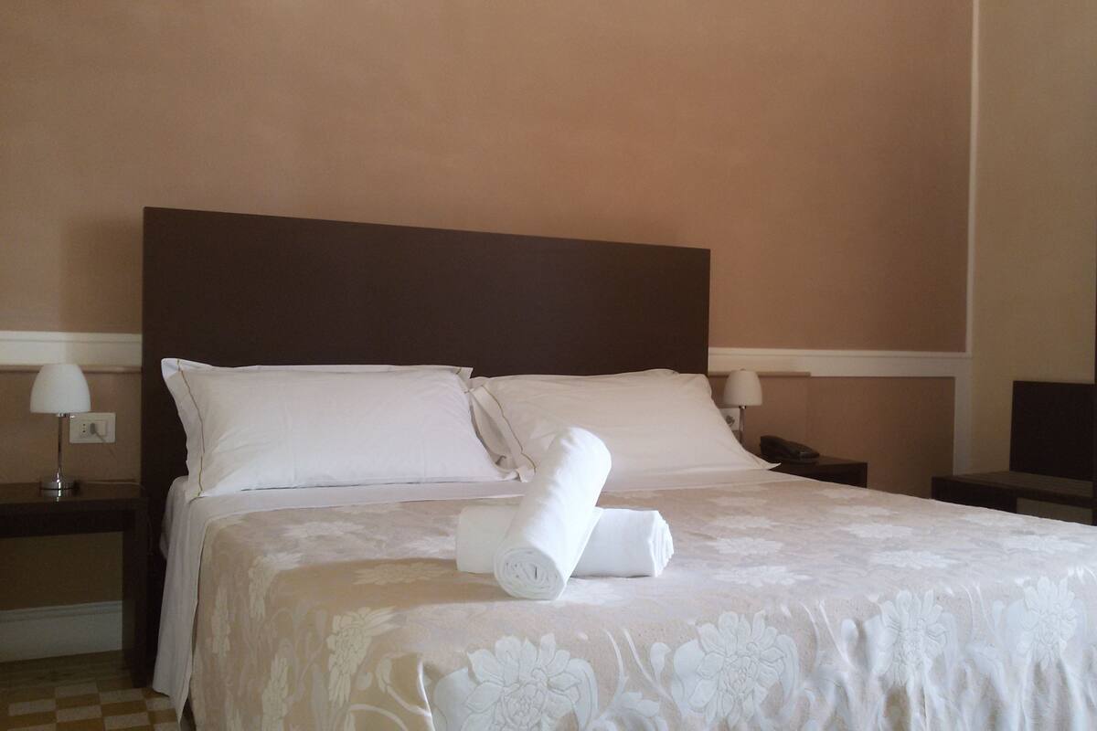 Een netjes opgemaakt bed met witte lakens en een gespreide laken met bloemenprint, twee witte kussens en een opgerolde handdoek op het bed.