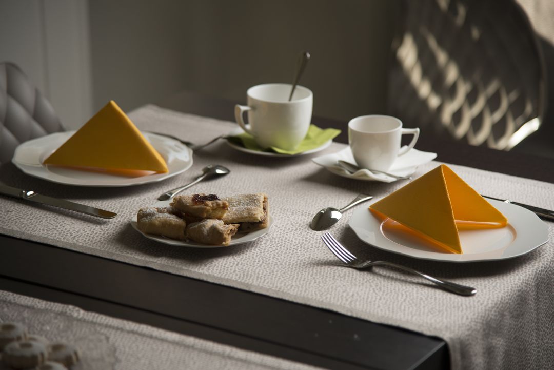 Una mesa dispuesta con dos tazas blancas, dos platos con objetos triangulares amarillos, un plato de pasteles y cubiertos.