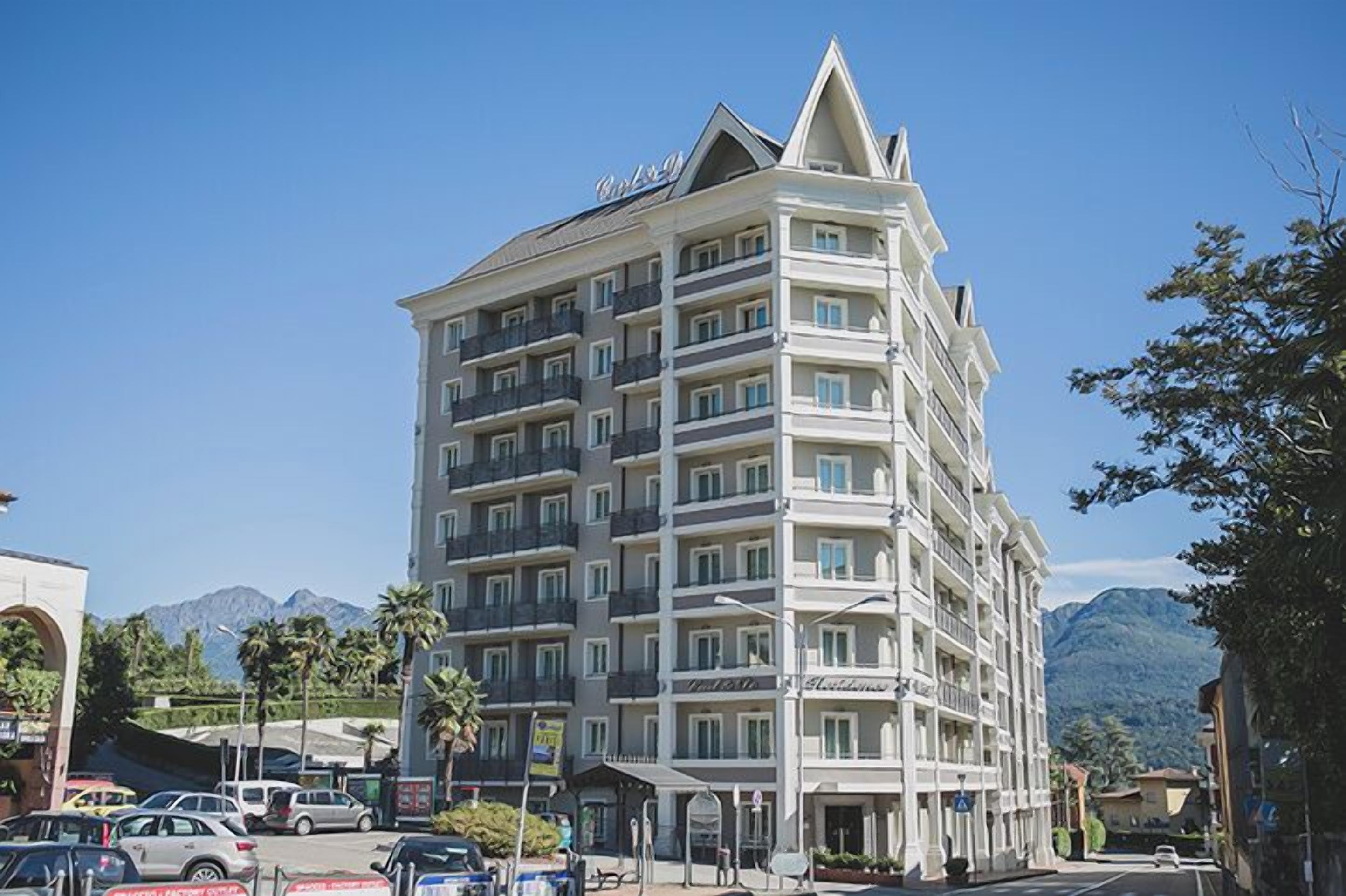 Un hotel de varios pisos con balcones, vistas despejadas a las montañas y un estacionamiento con automóviles.